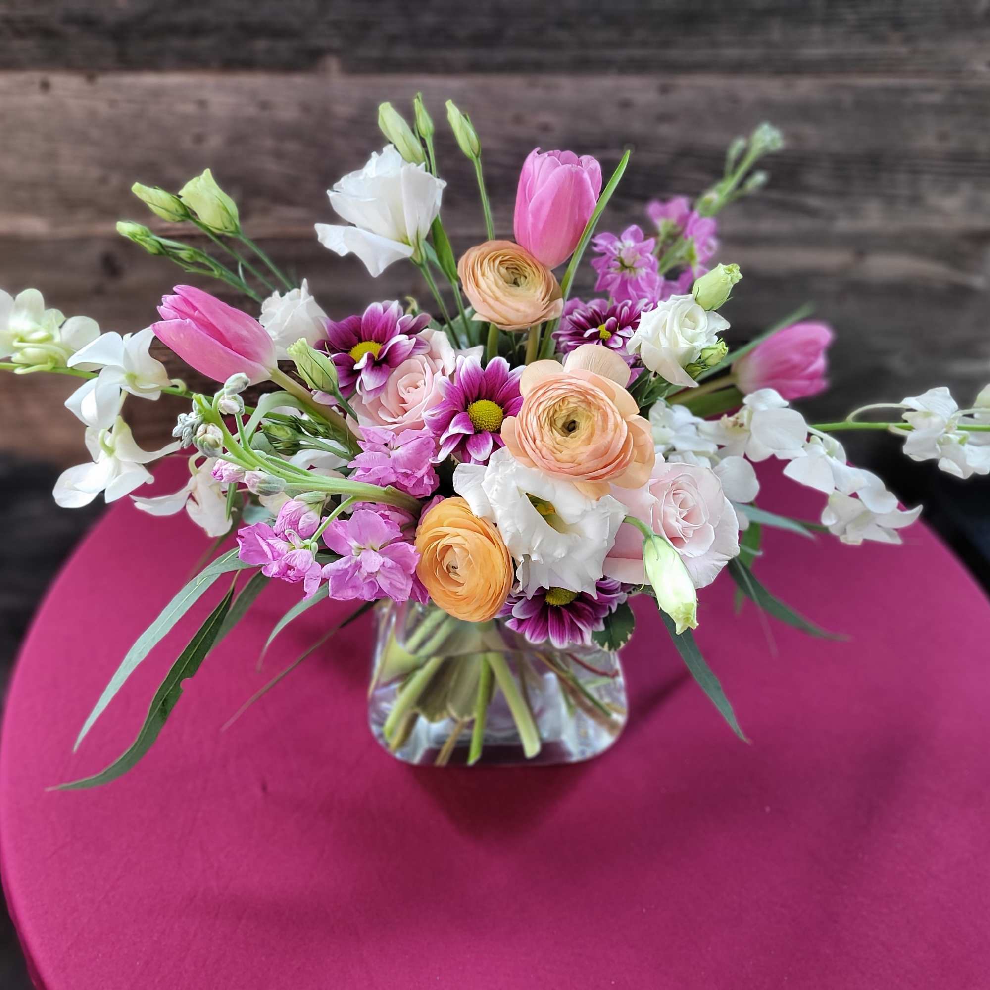 Low glass vase of pink tulips, peach ranunculus, white orchids, and mixed blooms on a magenta table.