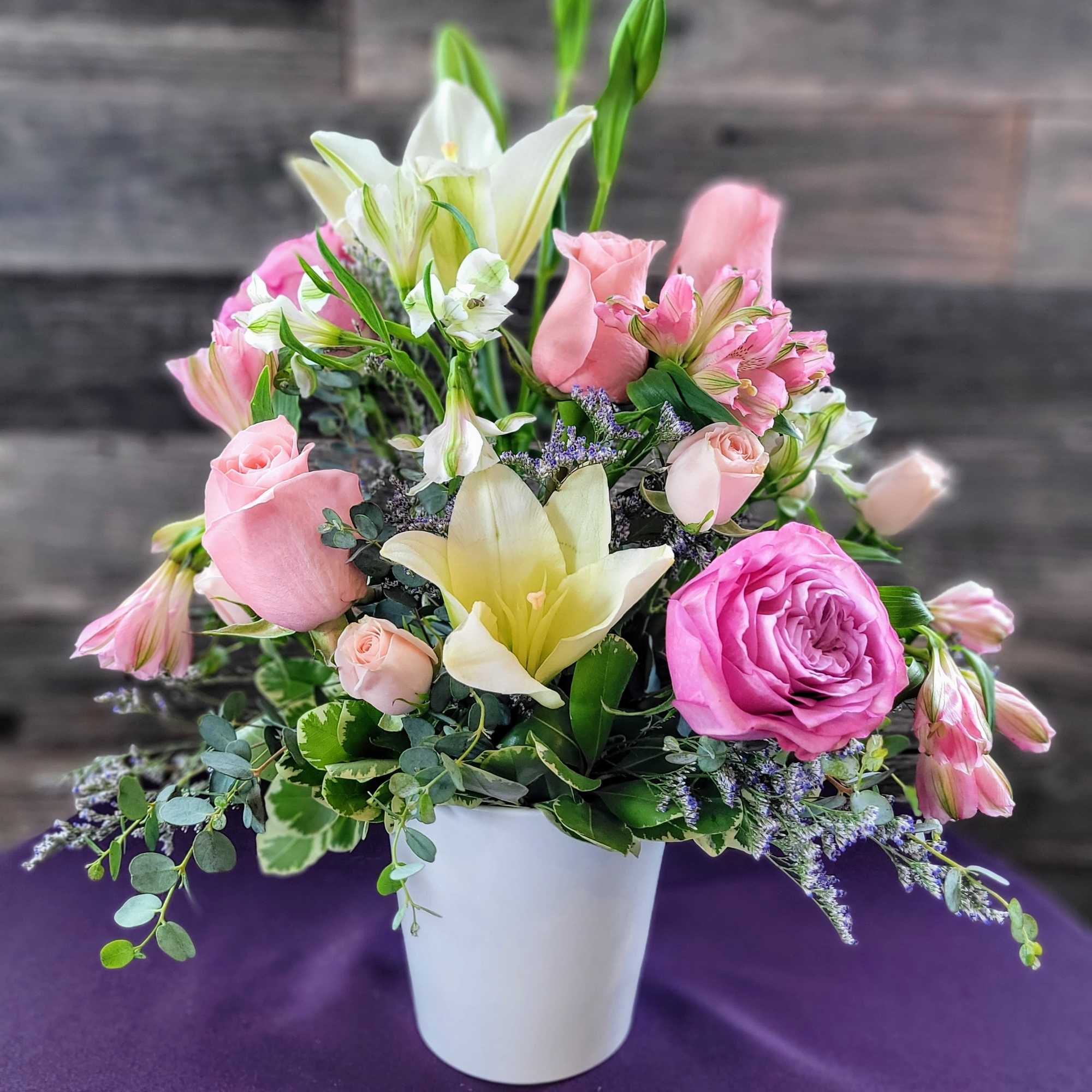 Pink roses and white lilies arranged in a white pot on a purple cloth.