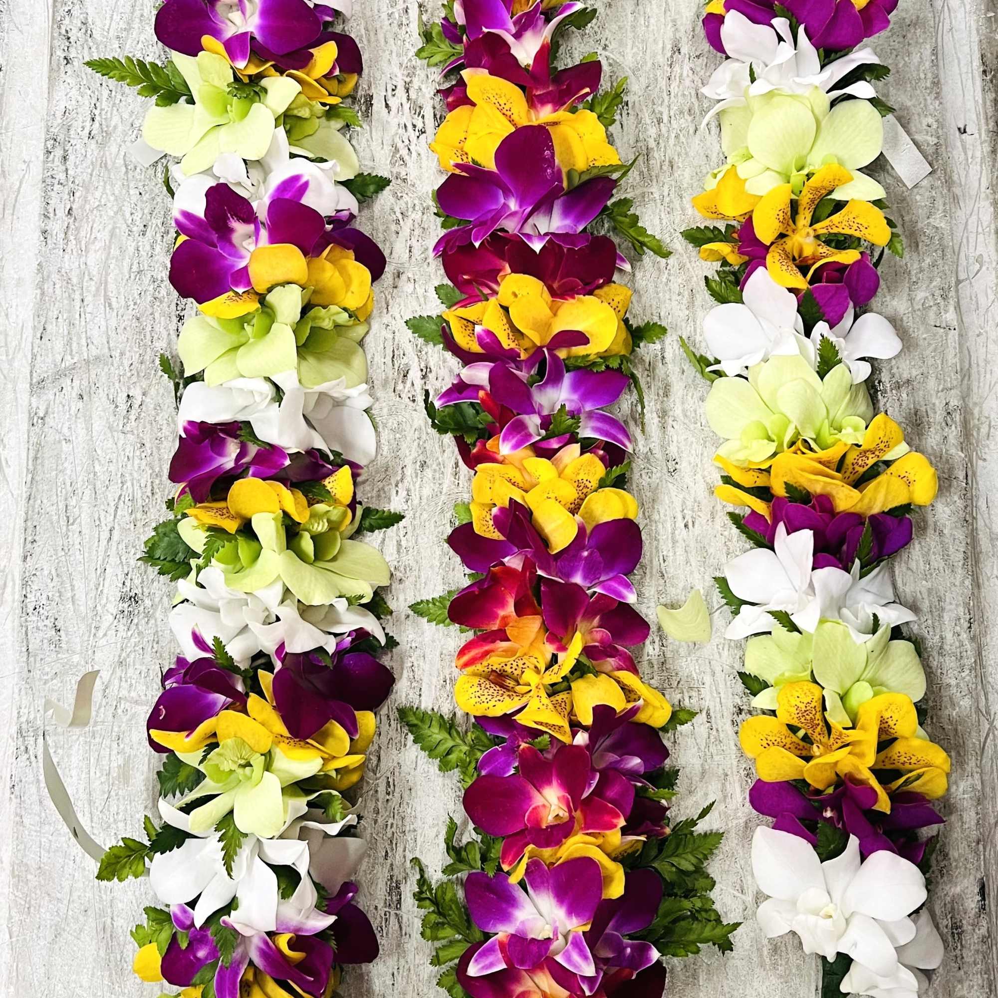 Three colorful orchid and lily floral leis in purple, yellow, white, and green with ribbon ties.