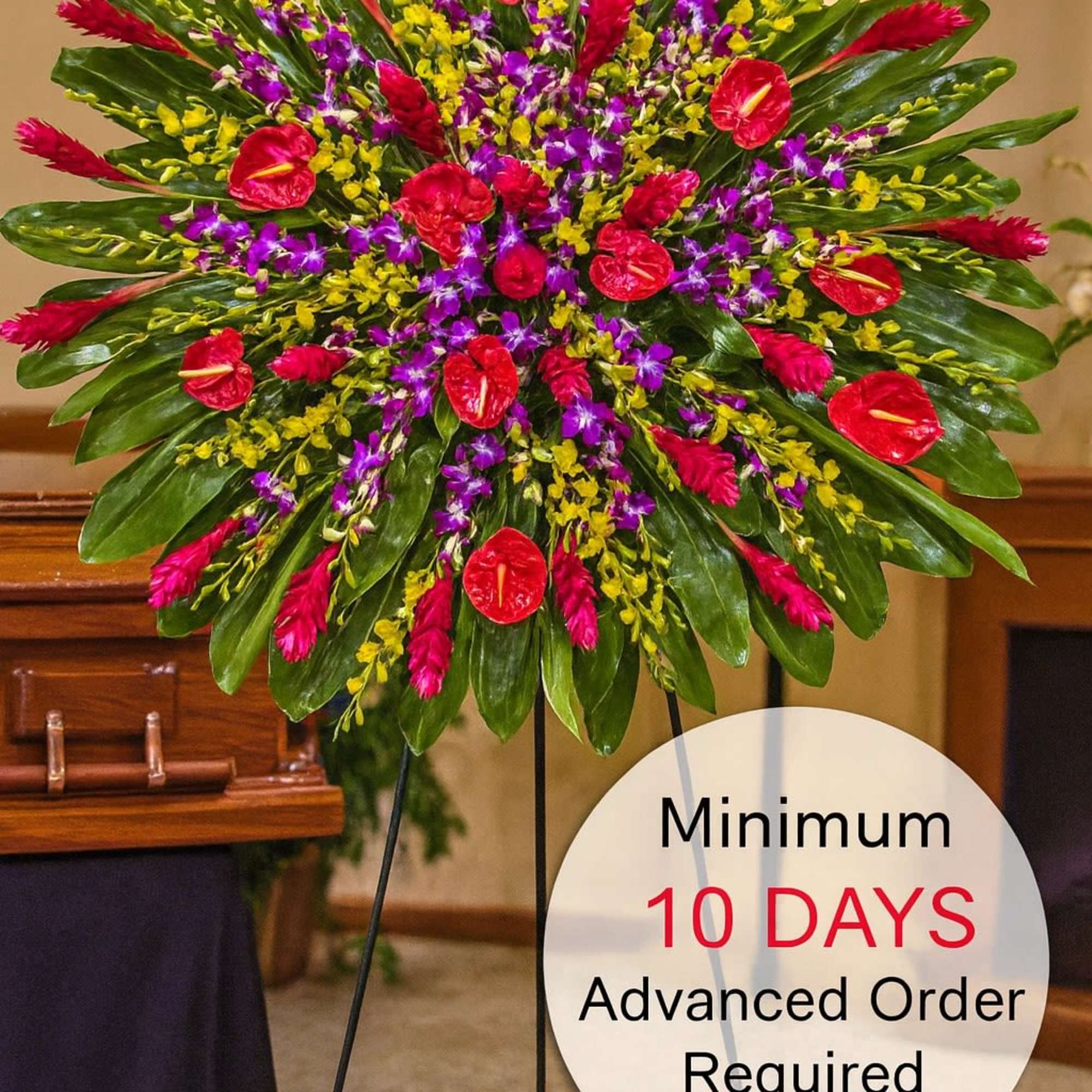 Large round tropical standing spray with red, purple, and yellow flowers on a metal easel in a funeral setting