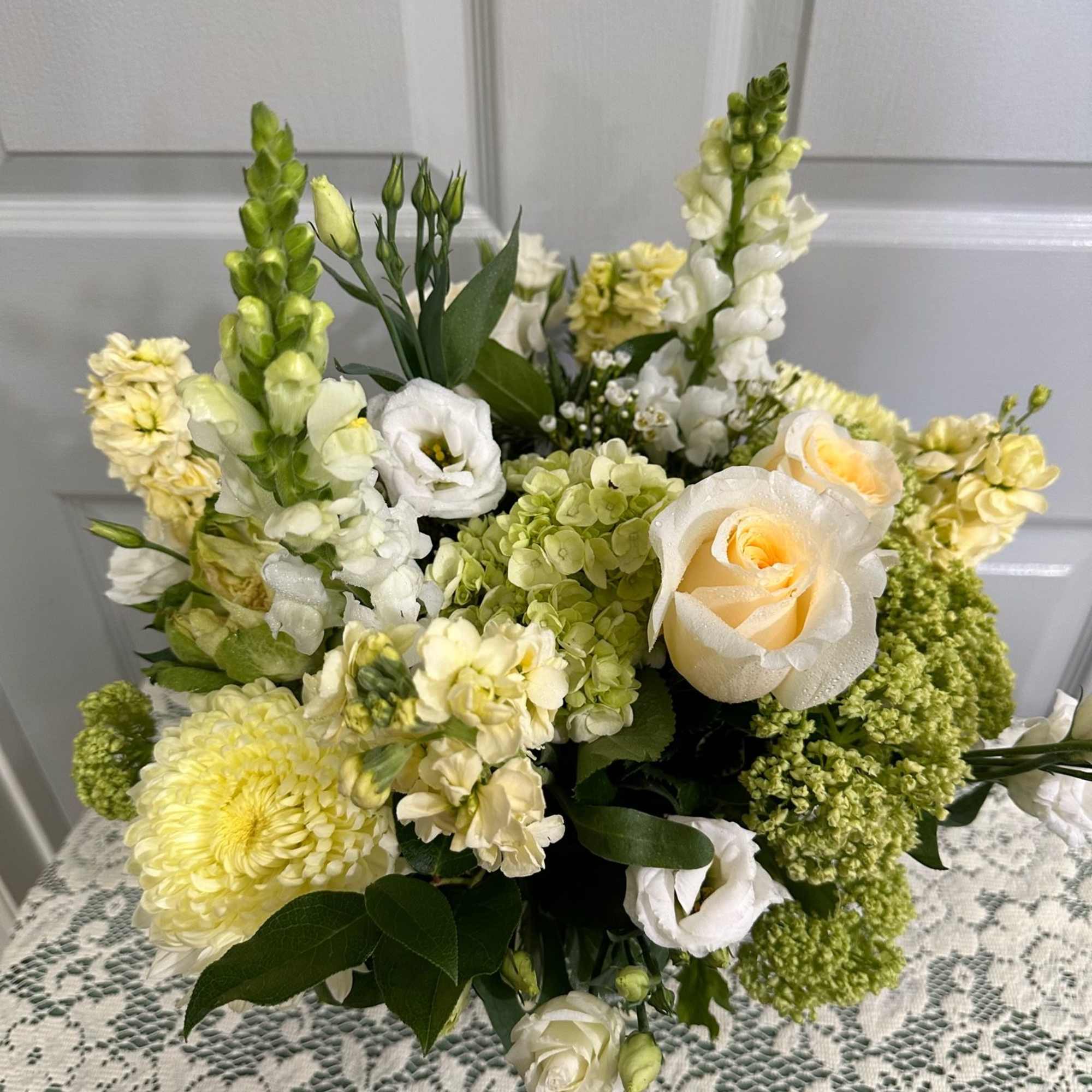 Cream and white floral arrangement with roses, lisianthus, snapdragons, hydrangeas, and mum on a lace table.