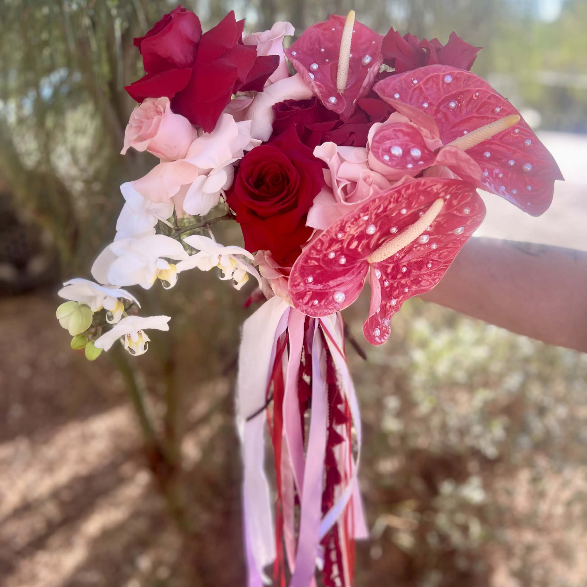 Handheld bouquet of red roses, pink anthuriums, and white orchids with ribbon tails