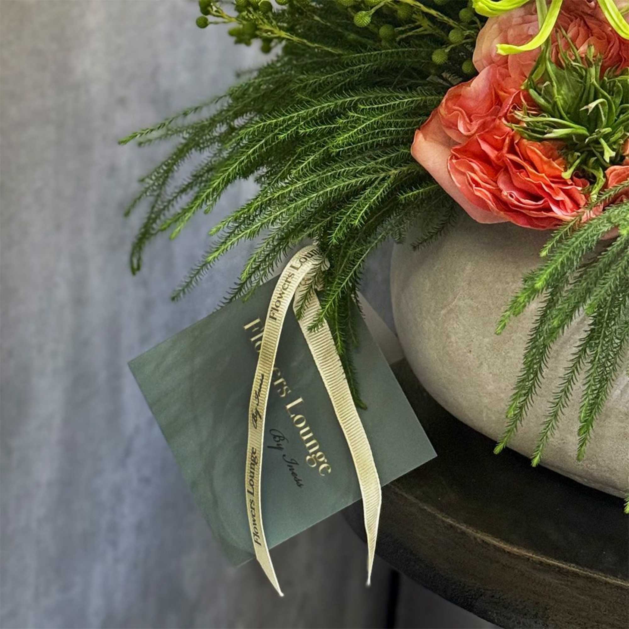Coral roses in a gray vase with trailing greenery and a ribboned card
