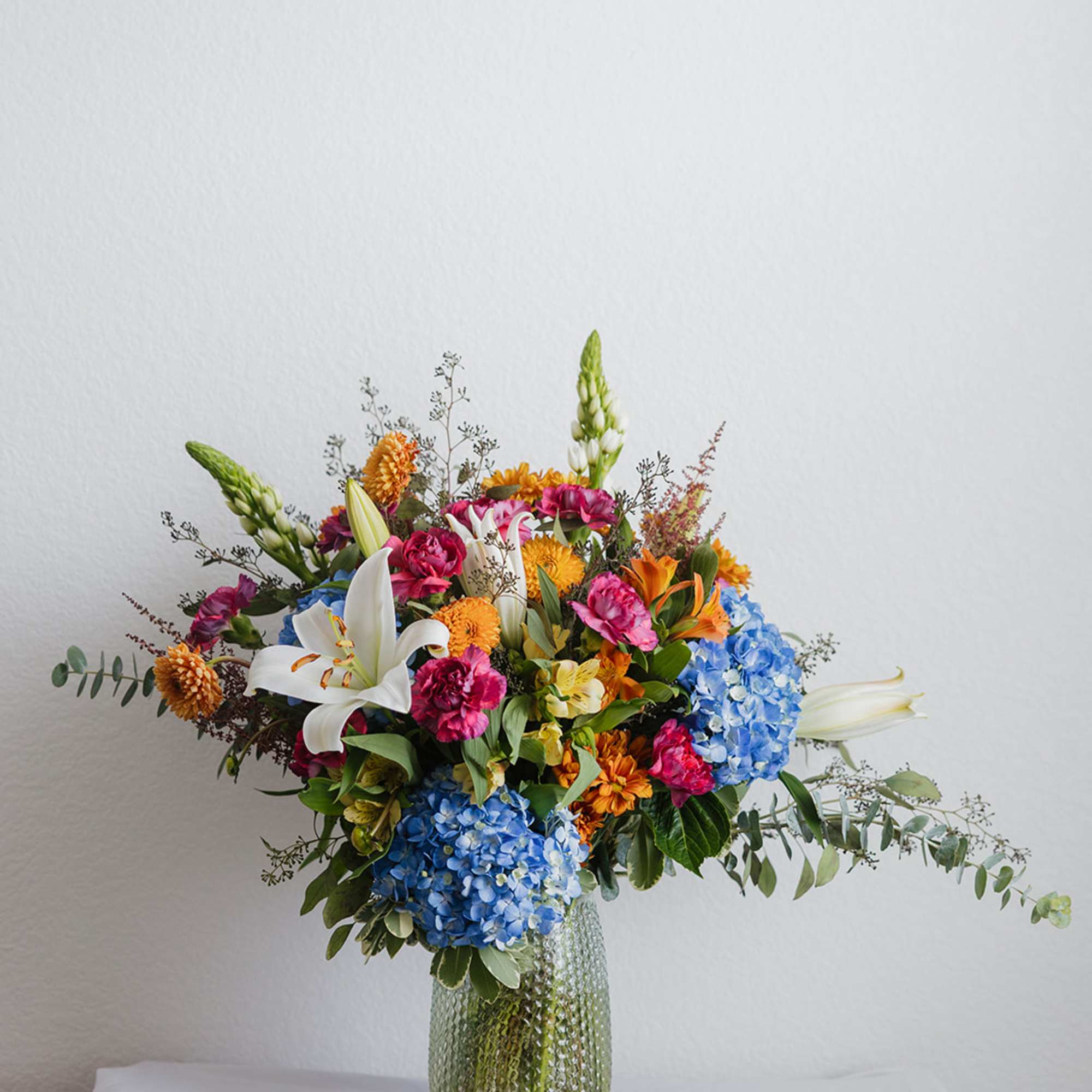 Mixed bouquet with blue hydrangeas, white lilies, and orange flowers in a glass vase