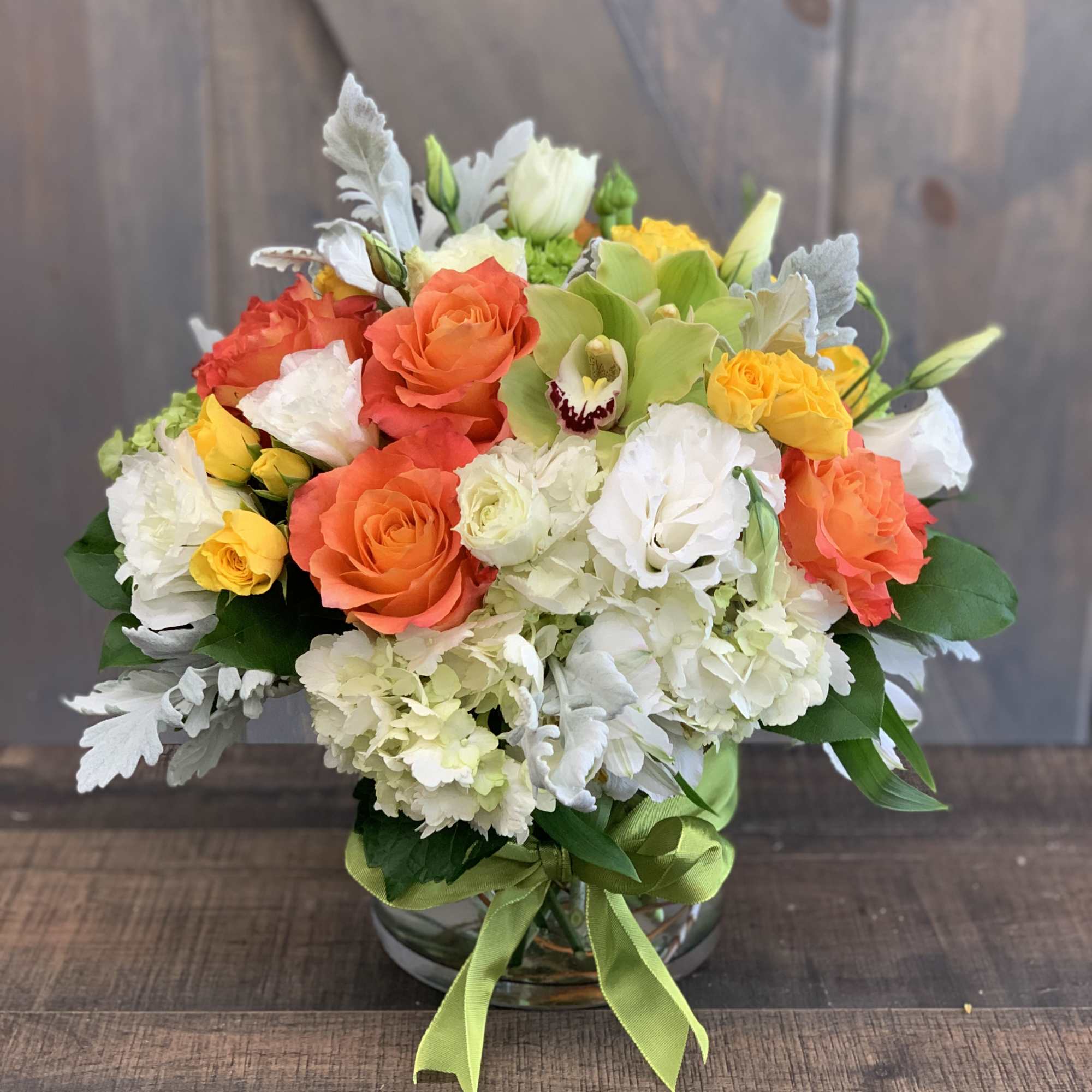 Mixed bouquet of orange, yellow, and white flowers in a glass vase with a green ribbon