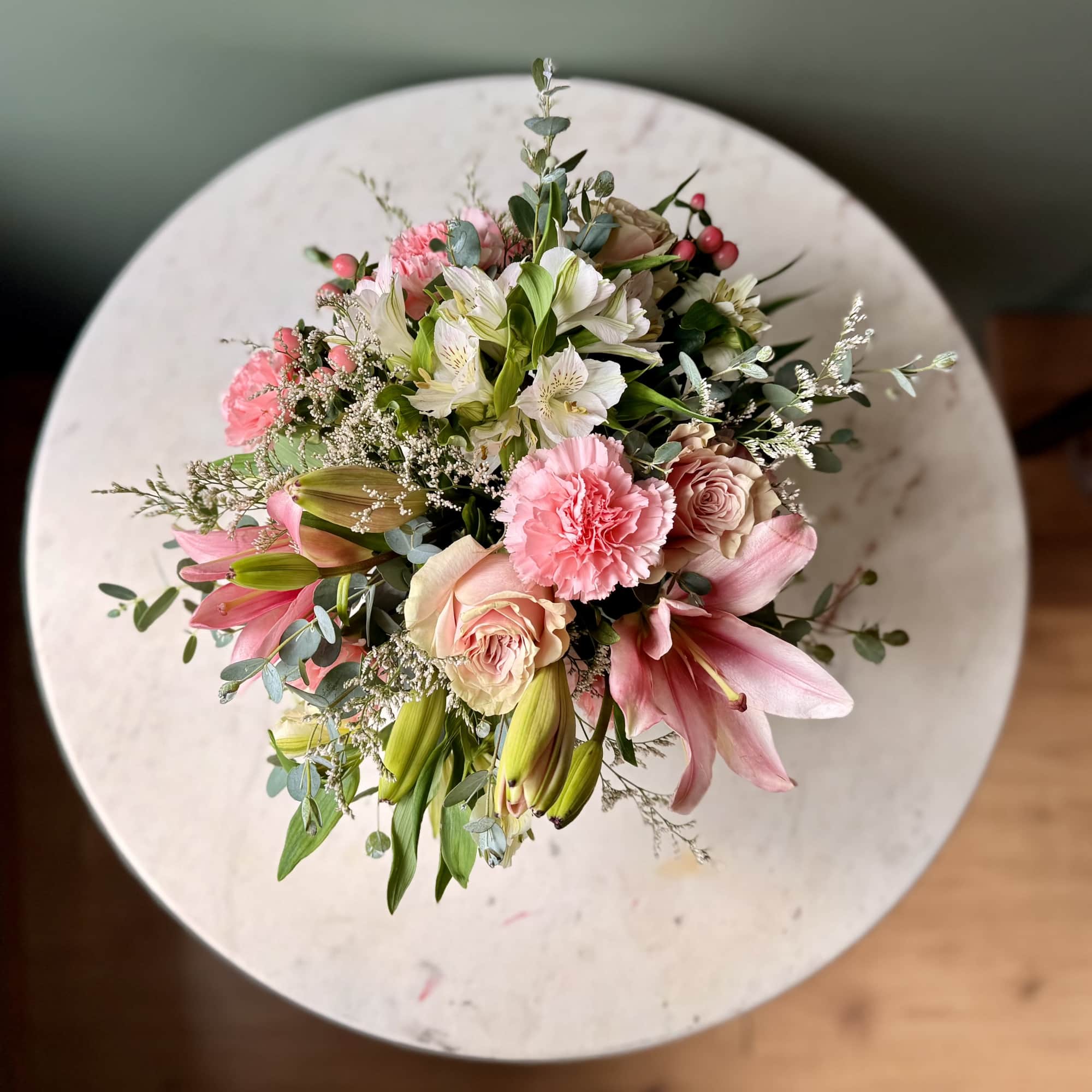 Top-down bouquet of pink lilies, roses, carnations, and white alstroemeria on a round table.