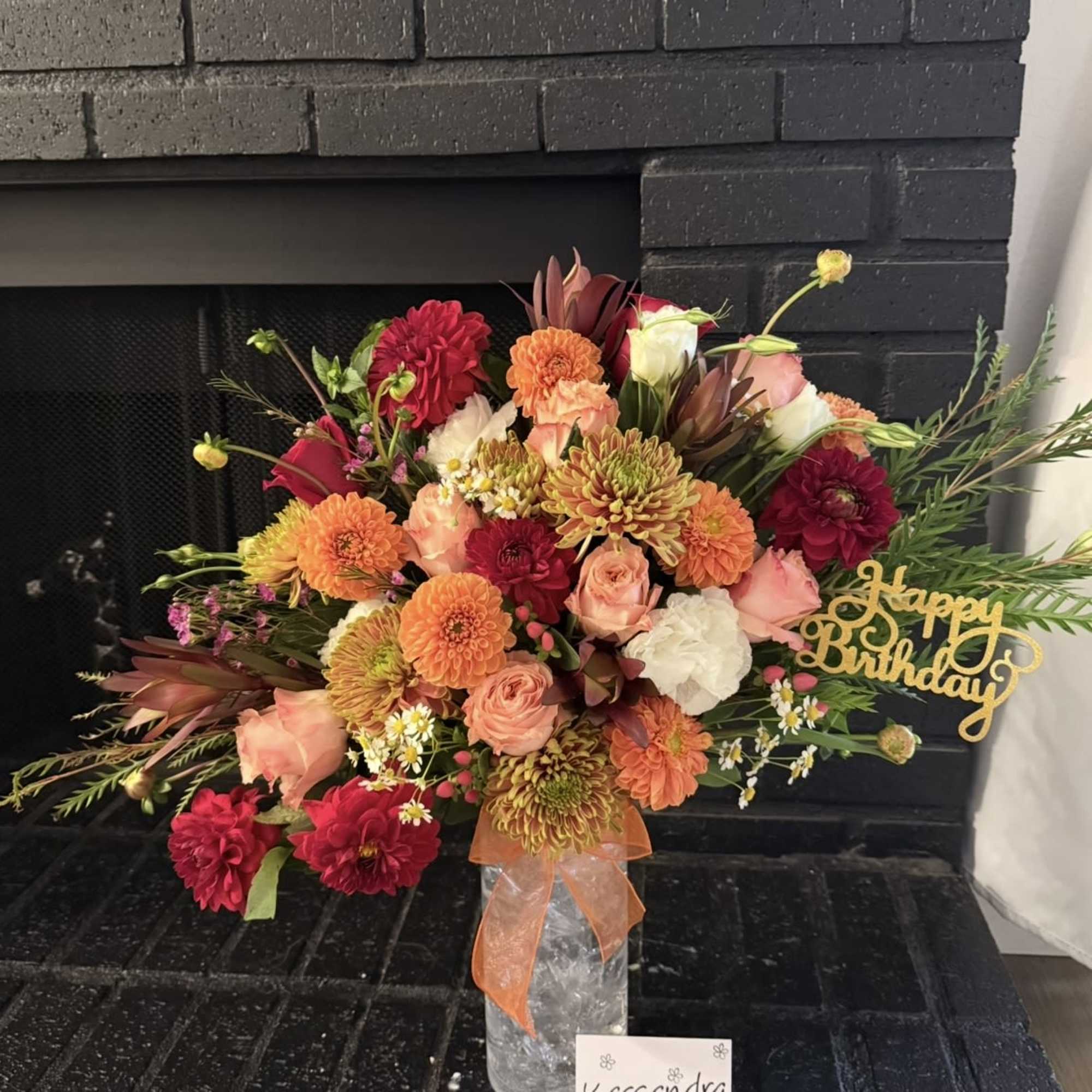 Mixed bouquet of pink, orange, and red flowers in a clear vase with a birthday sign