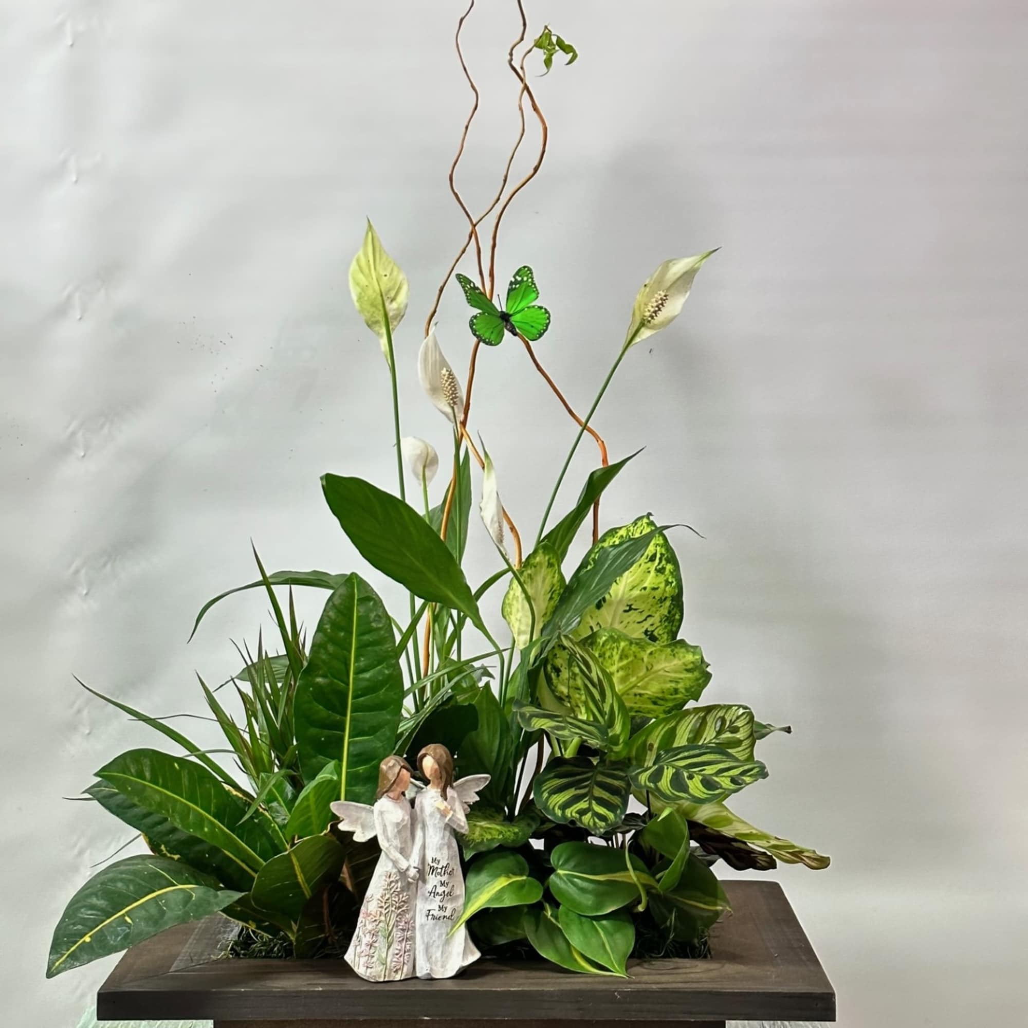 Tropical plant arrangement with white peace lilies and a figurine on a dark pedestal