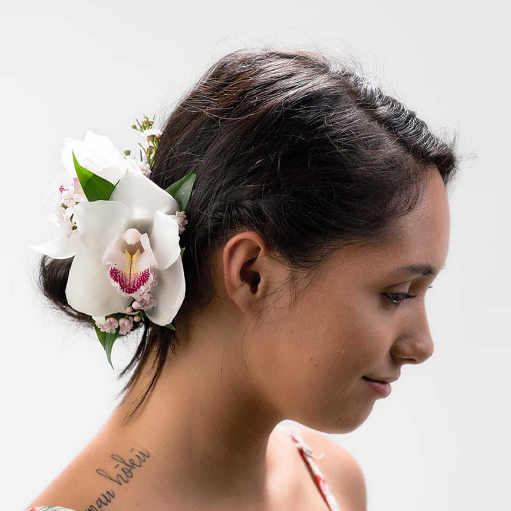 Floral hairpiece with a large white orchid and small pink blooms worn in dark hair styled in a low bun.