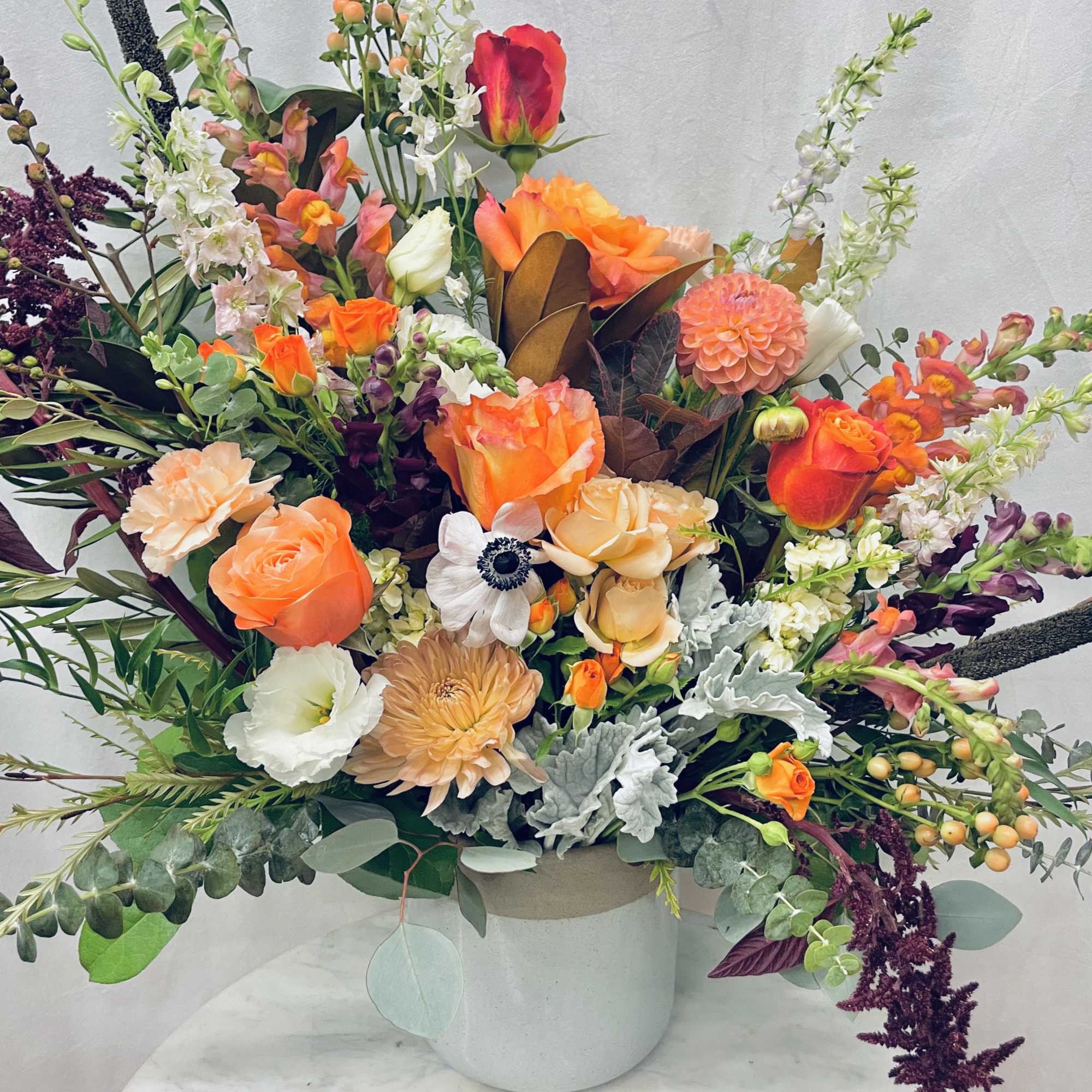 Large mixed floral arrangement in a white vase with orange, peach, white, and purple blooms