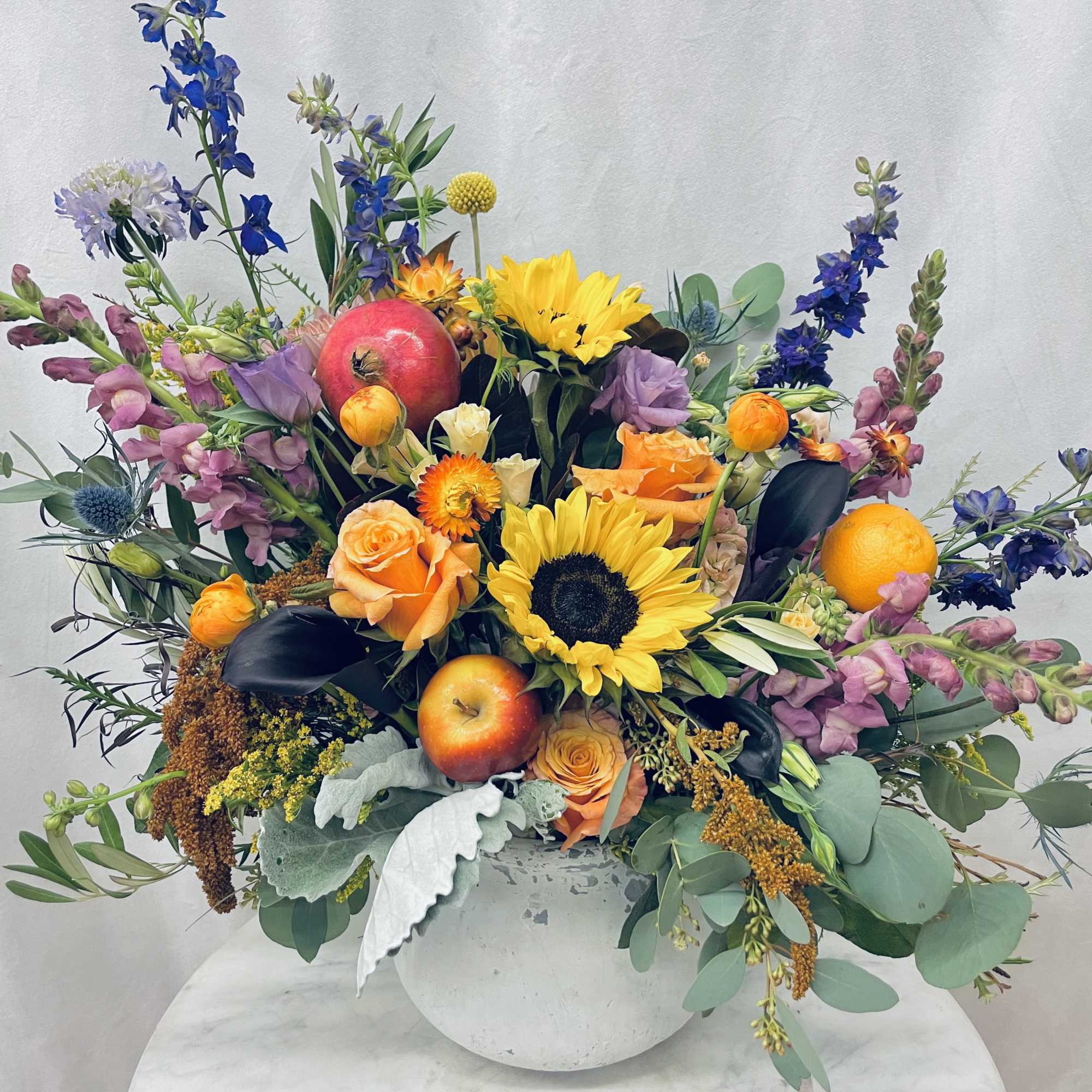Mixed bouquet with sunflowers, roses, snapdragons, and fruit in a white vase