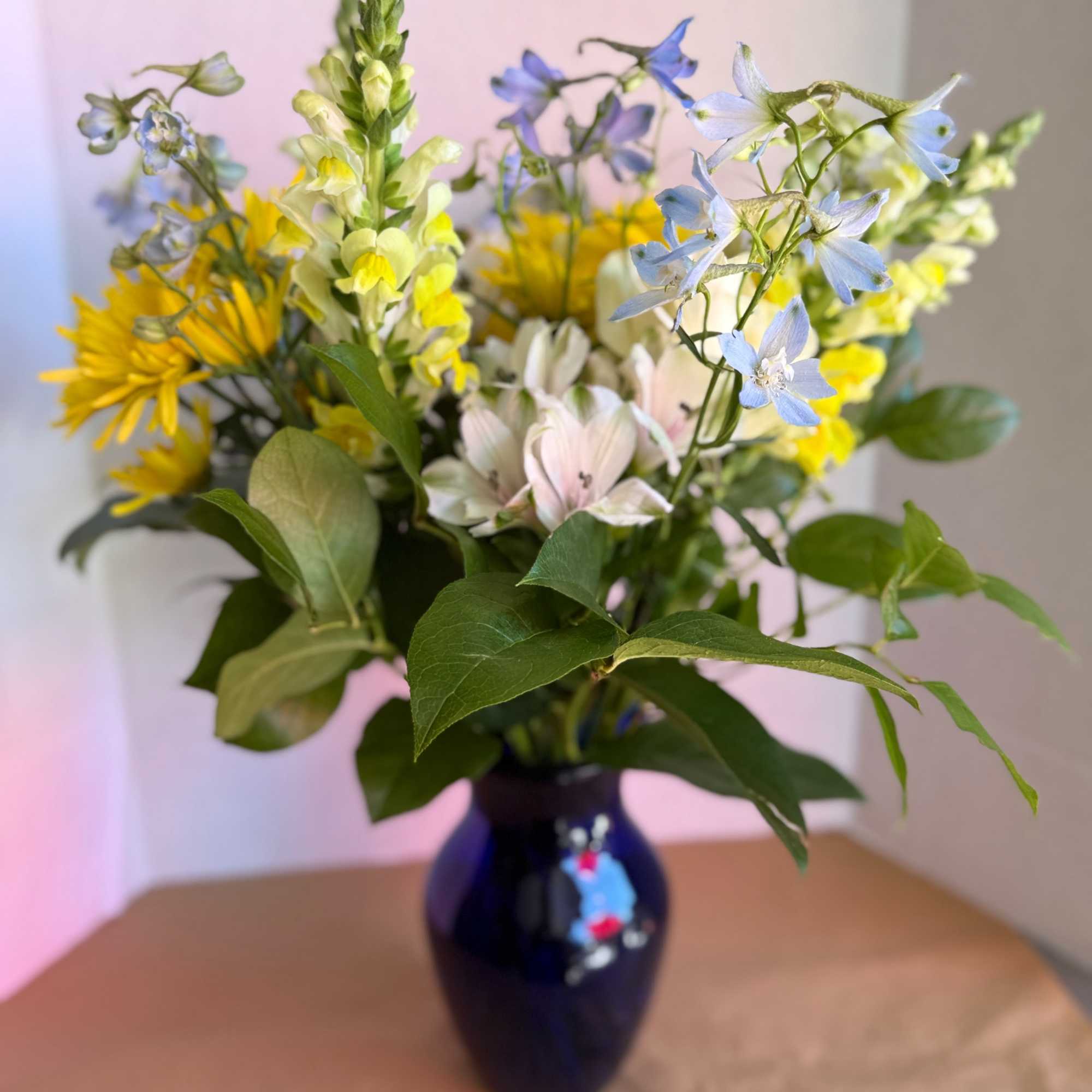 Mixed bouquet of yellow, blue, and pale pink flowers in a dark vase