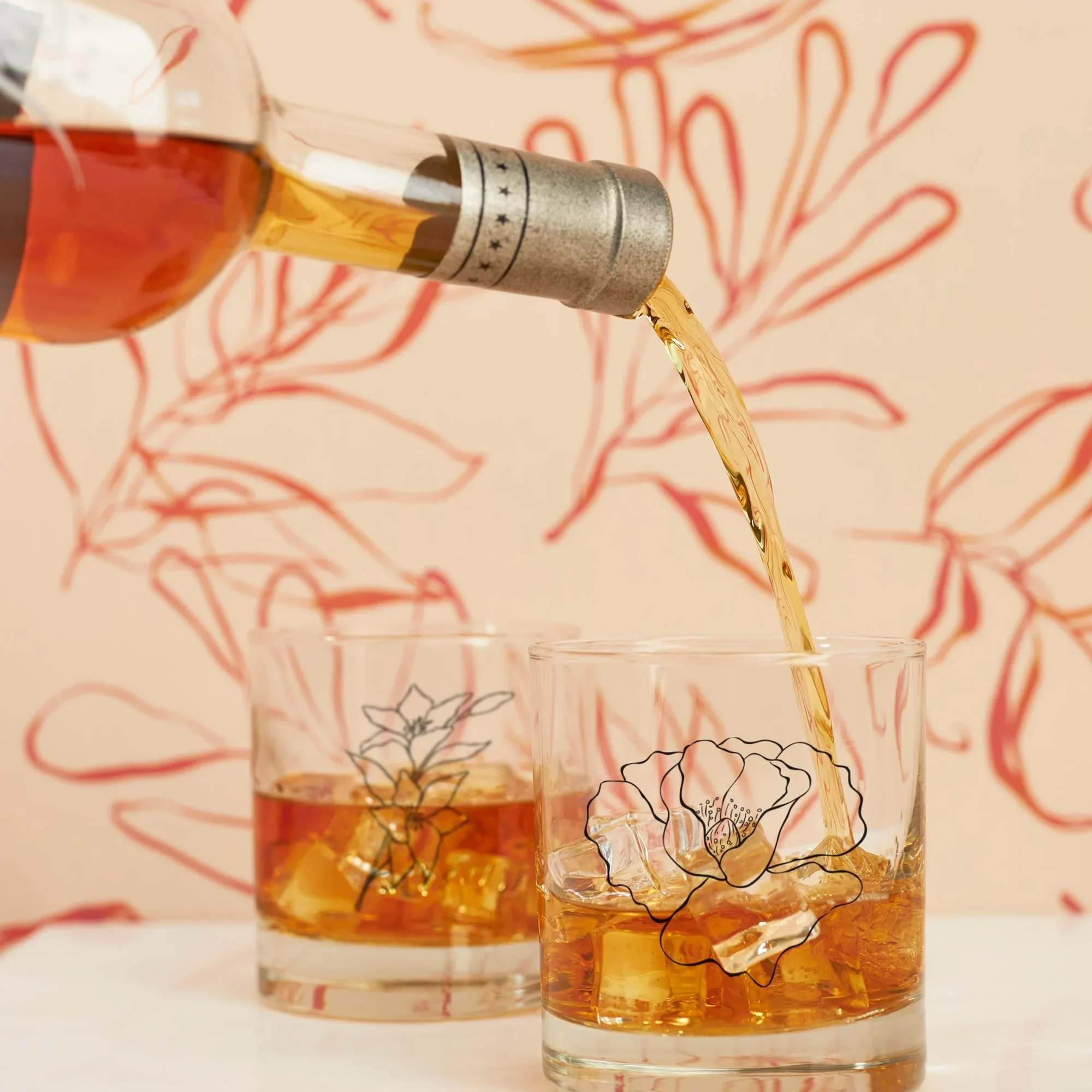 A bottle pours amber liquid into two floral-printed low glasses