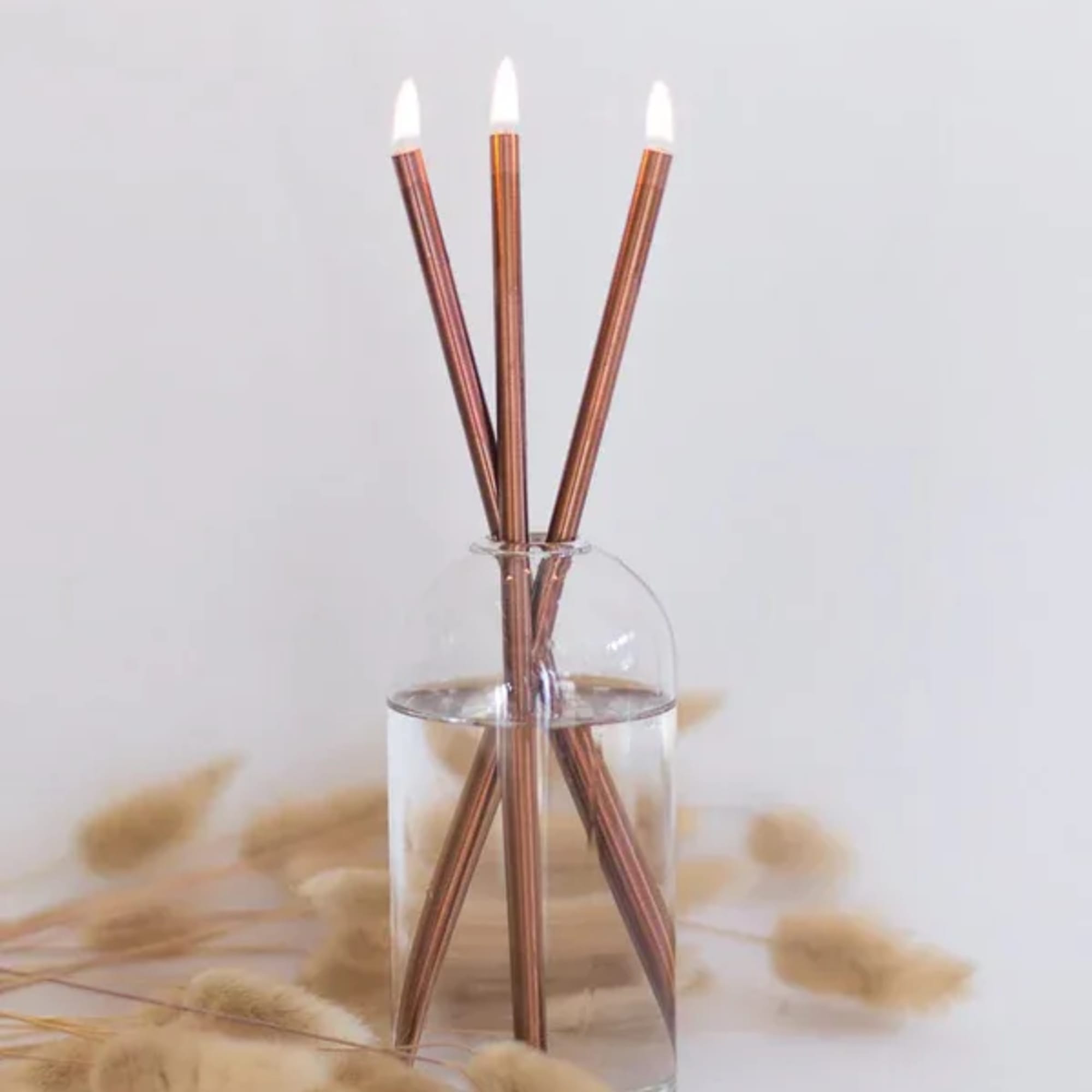 Three copper-toned taper candles in a clear glass vase