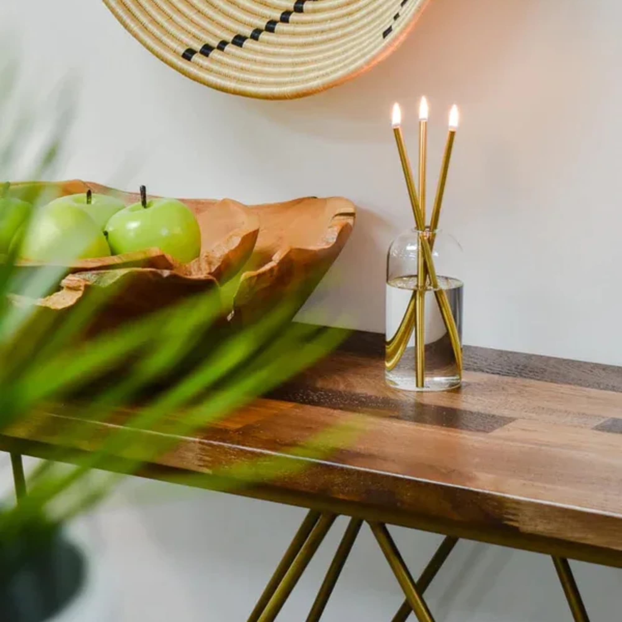 Glass vase with three lit taper candles on a wooden table beside green apples