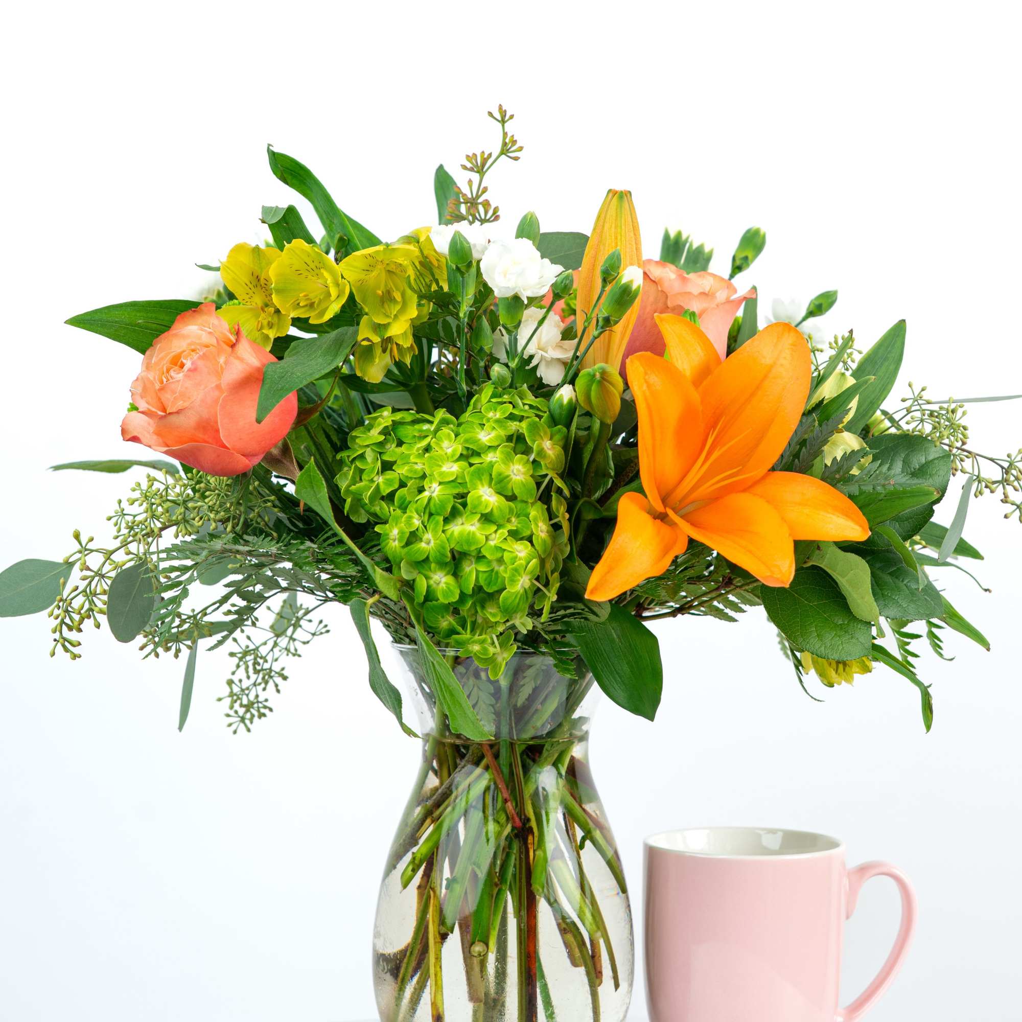 Mixed bouquet with orange lilies, peach roses, green hydrangea in a clear vase beside a pink mug