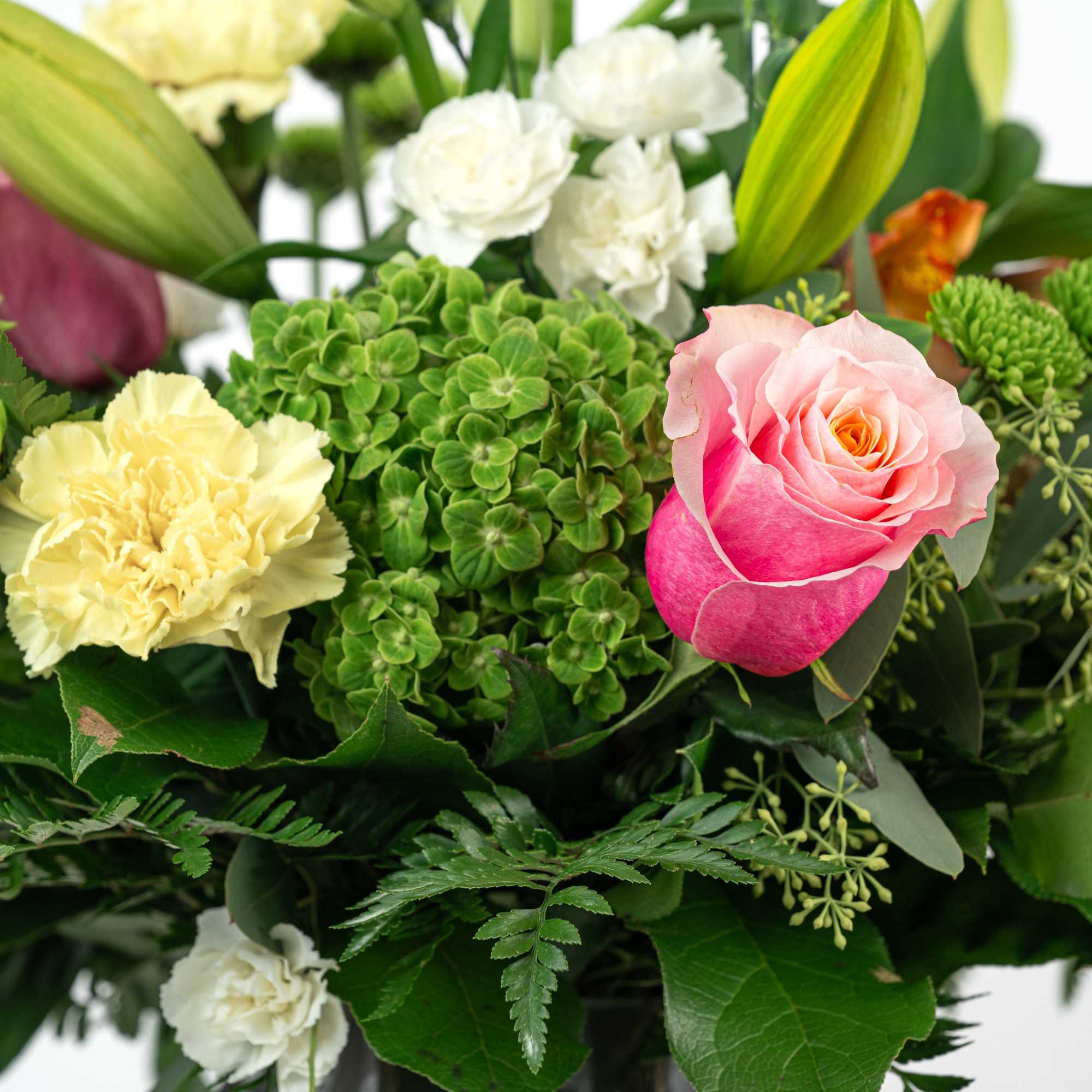Mixed bouquet with pink rose, yellow carnations, white blooms, green hydrangea, and lily buds in a glass vase
