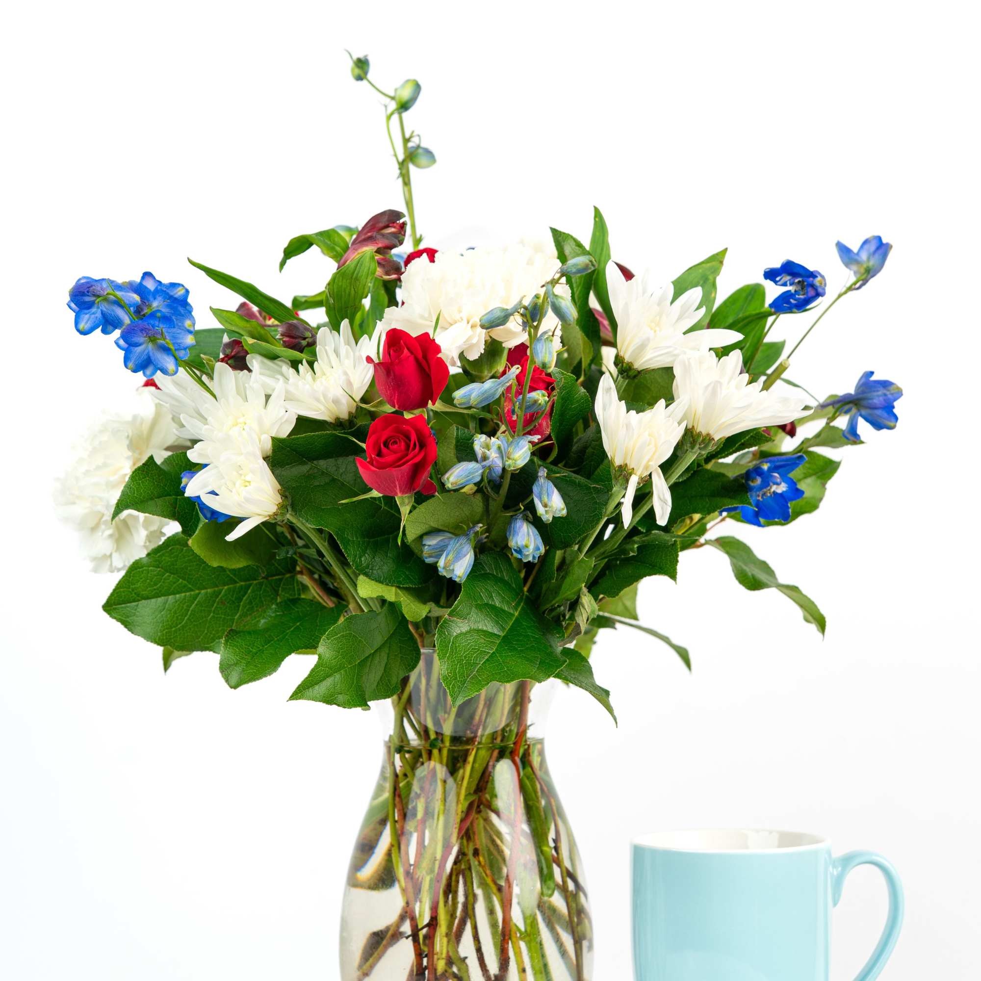 Mixed white, red, and blue flower arrangement in a clear glass vase beside a light blue mug.