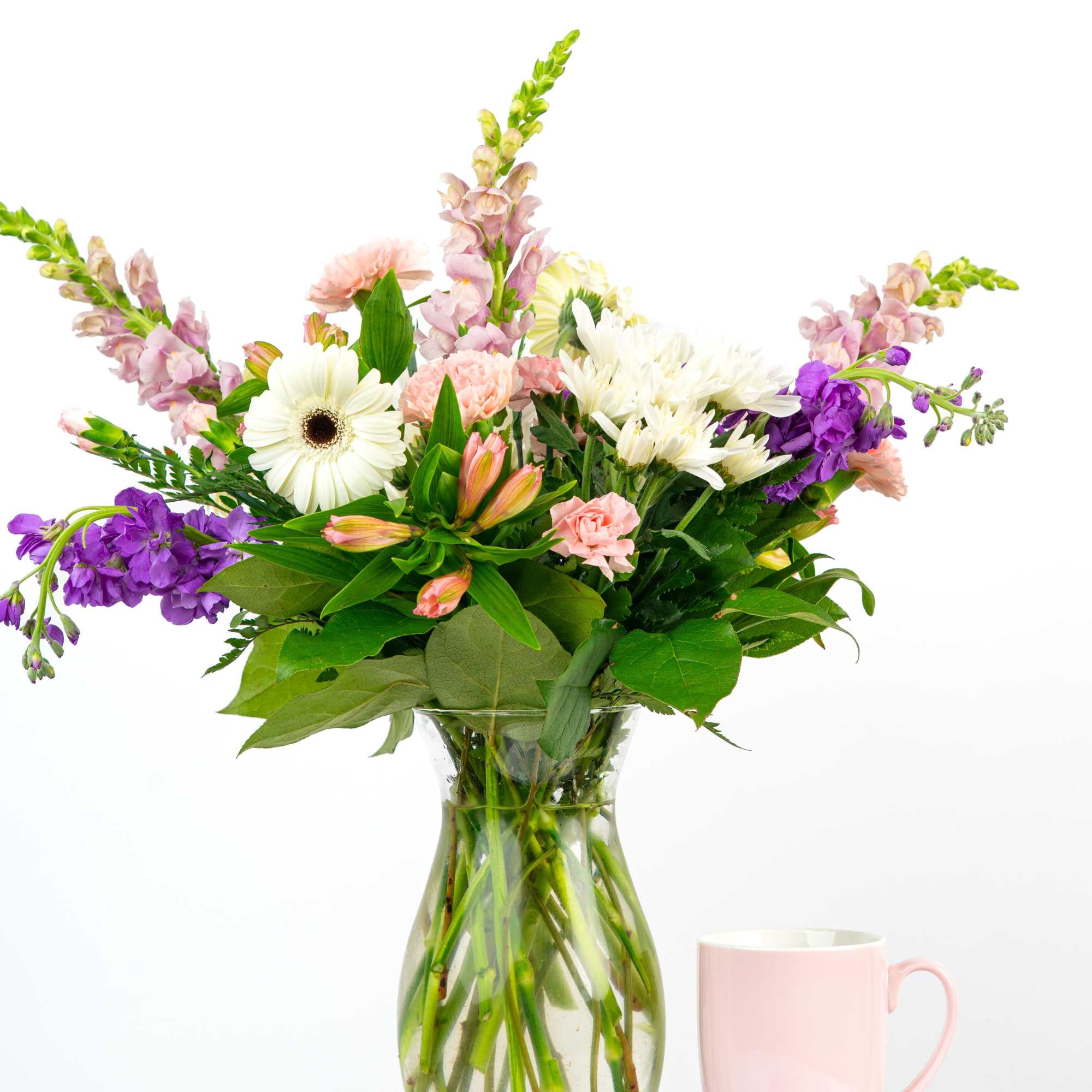 Mixed pink, white, and purple flower arrangement in a clear glass vase beside a pale pink mug.