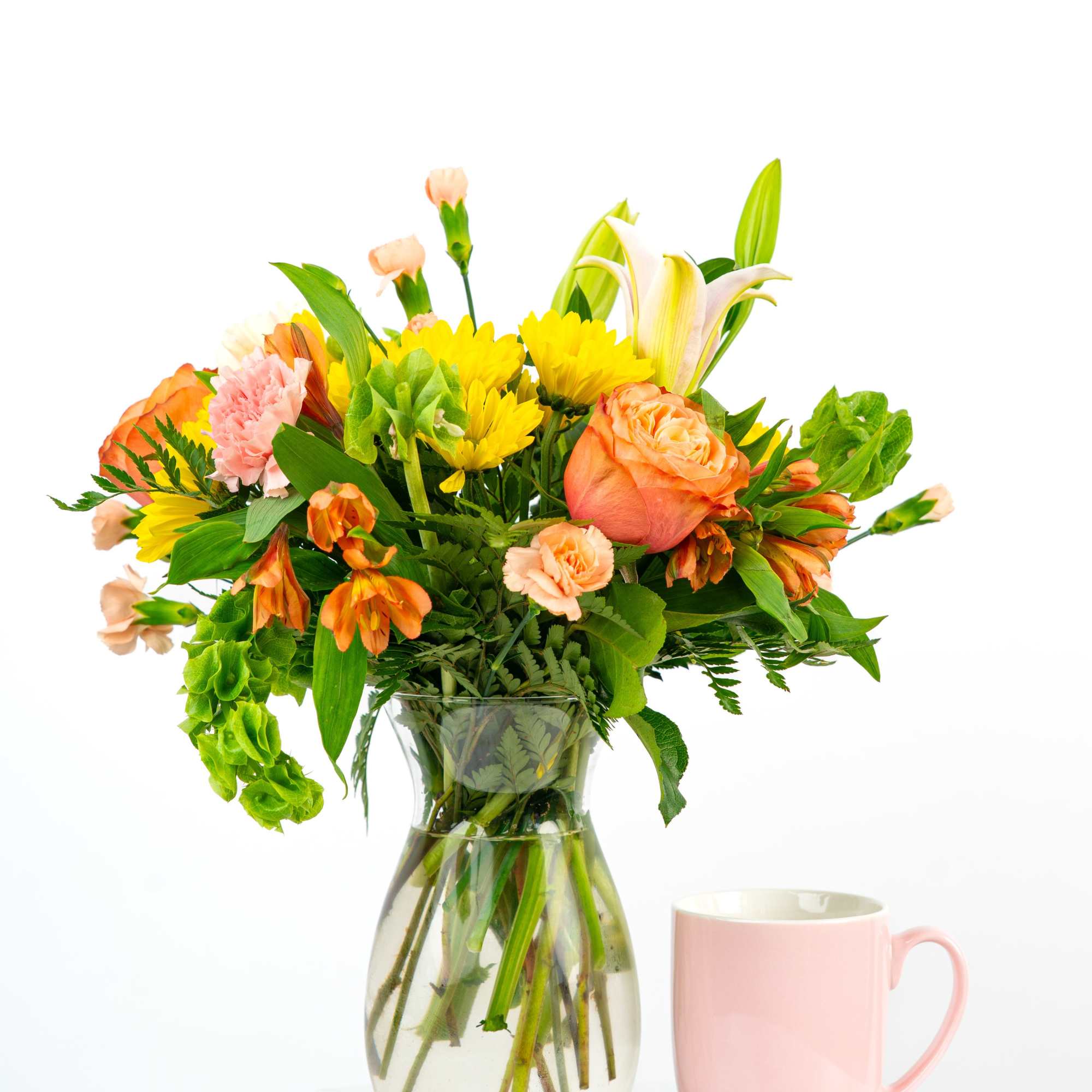 Mixed orange, yellow, and pink flower bouquet in a clear glass vase beside a pale pink mug