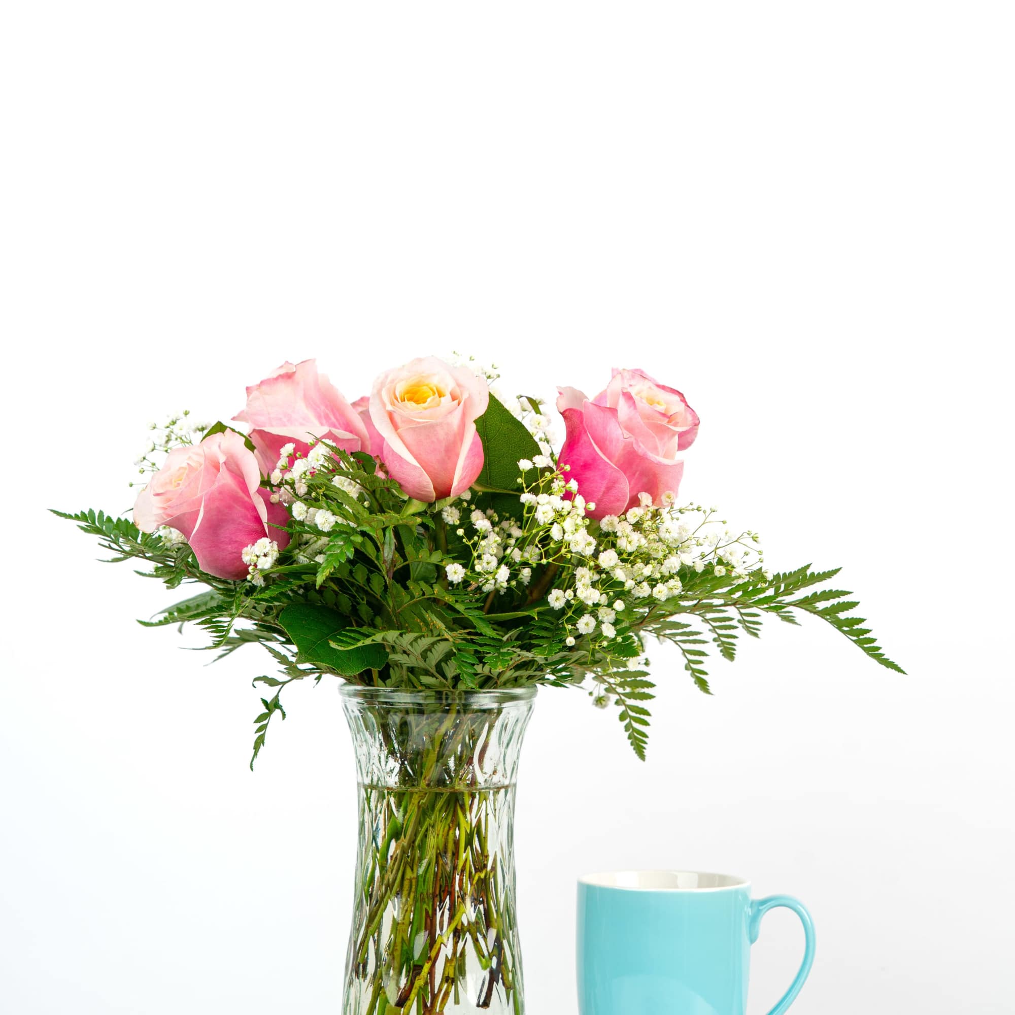 Pink roses and white filler flowers in a clear glass vase beside a light blue mug