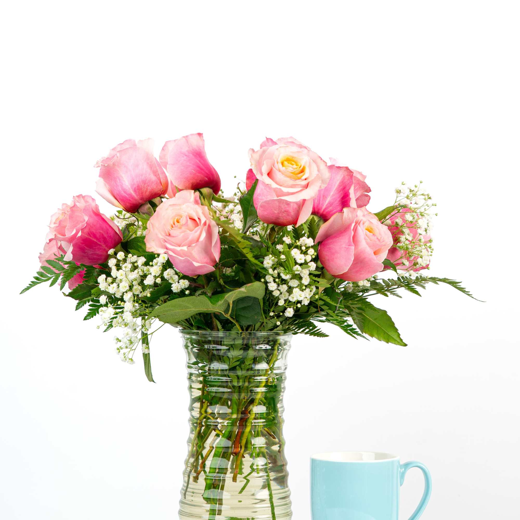 Tall clear vase of pink roses with white filler flowers beside a light blue mug