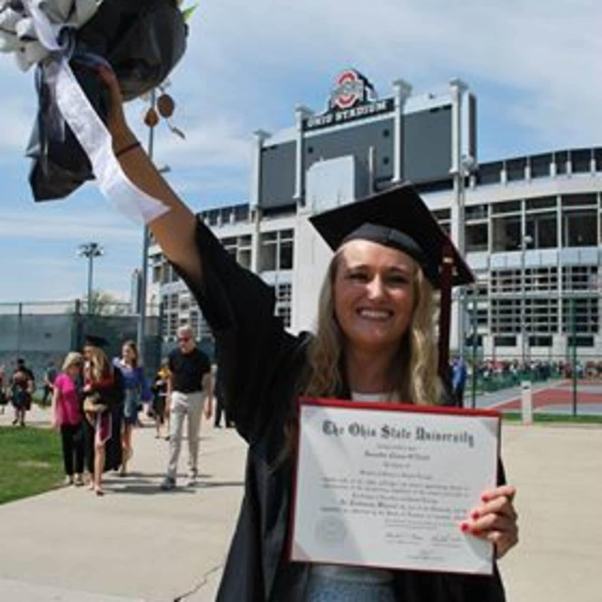 Graduate in cap and gown holding a bouquet and diploma outside a stadium