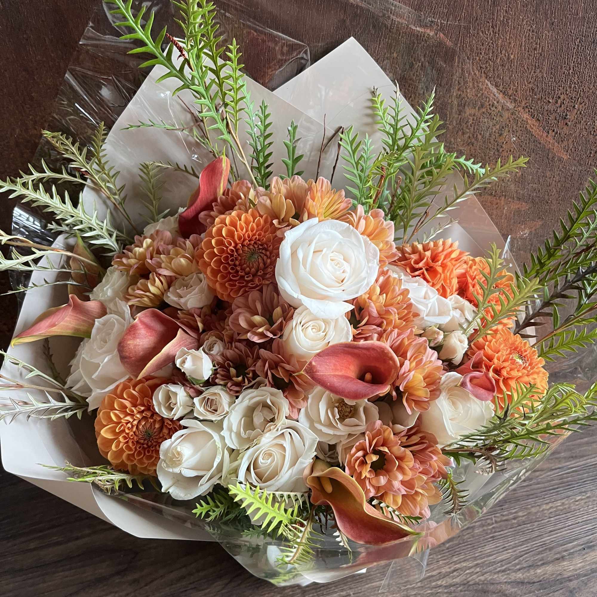 Bouquet of white roses, orange blooms, and pink calla lilies wrapped in clear plastic