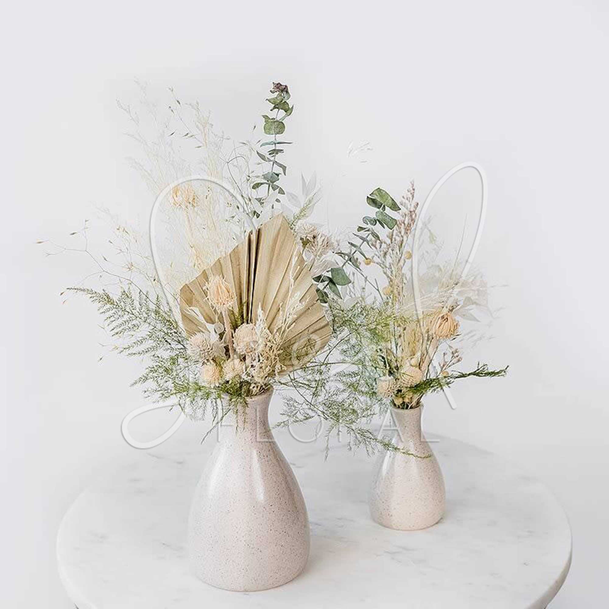 Two dried floral arrangements in cream vases on a round table