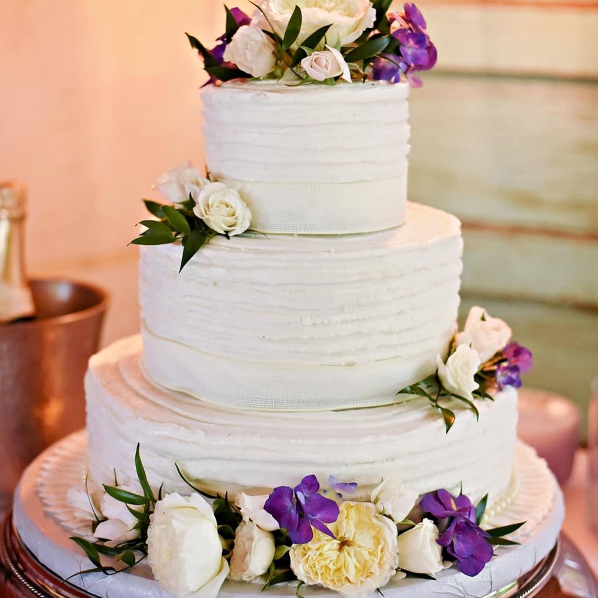Three-tier white cake decorated with white and purple flowers on a silver stand.