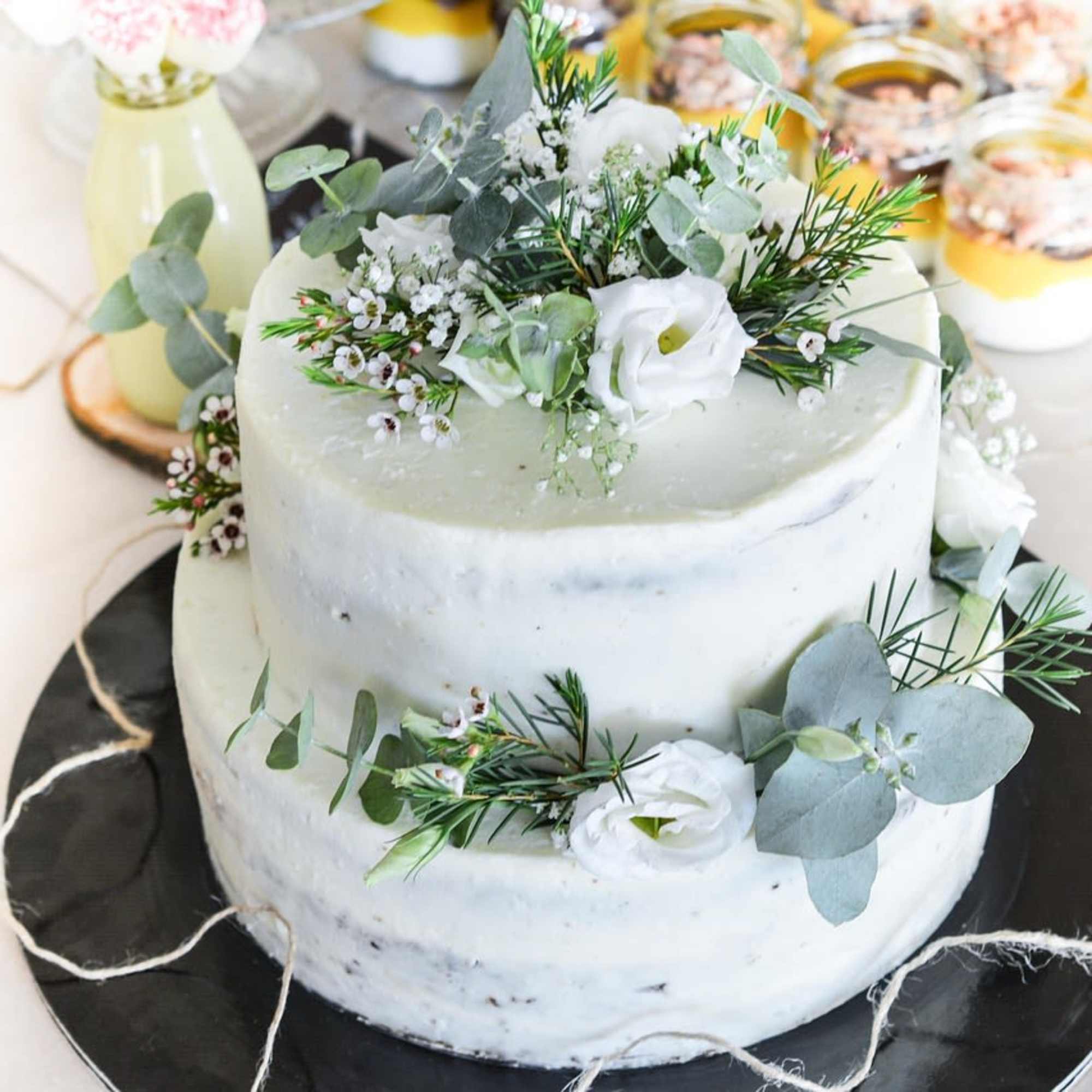 Two-tier white cake decorated with small white flowers and sprigs of greenery on a black stand.