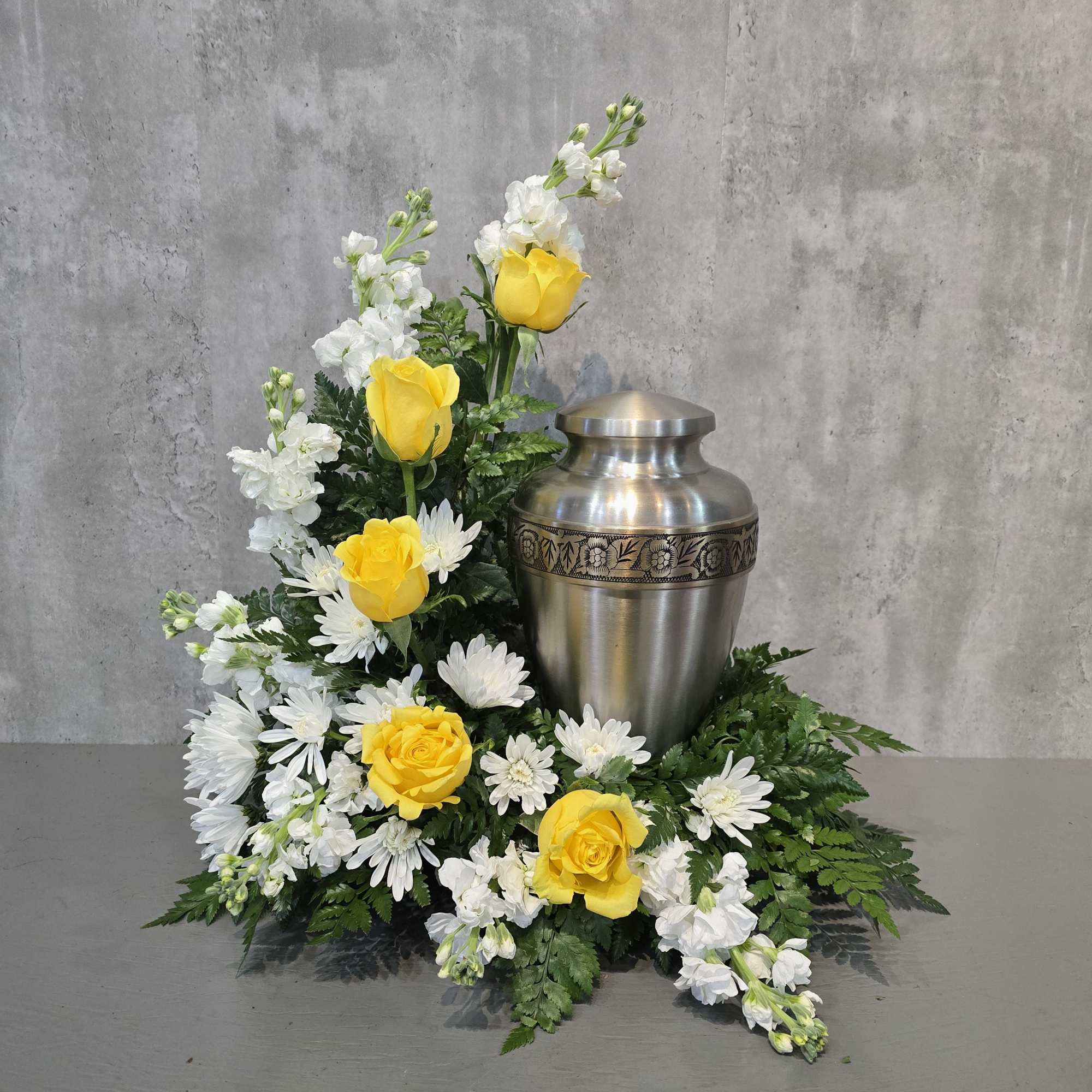Yellow roses and white flowers arranged around a silver urn.