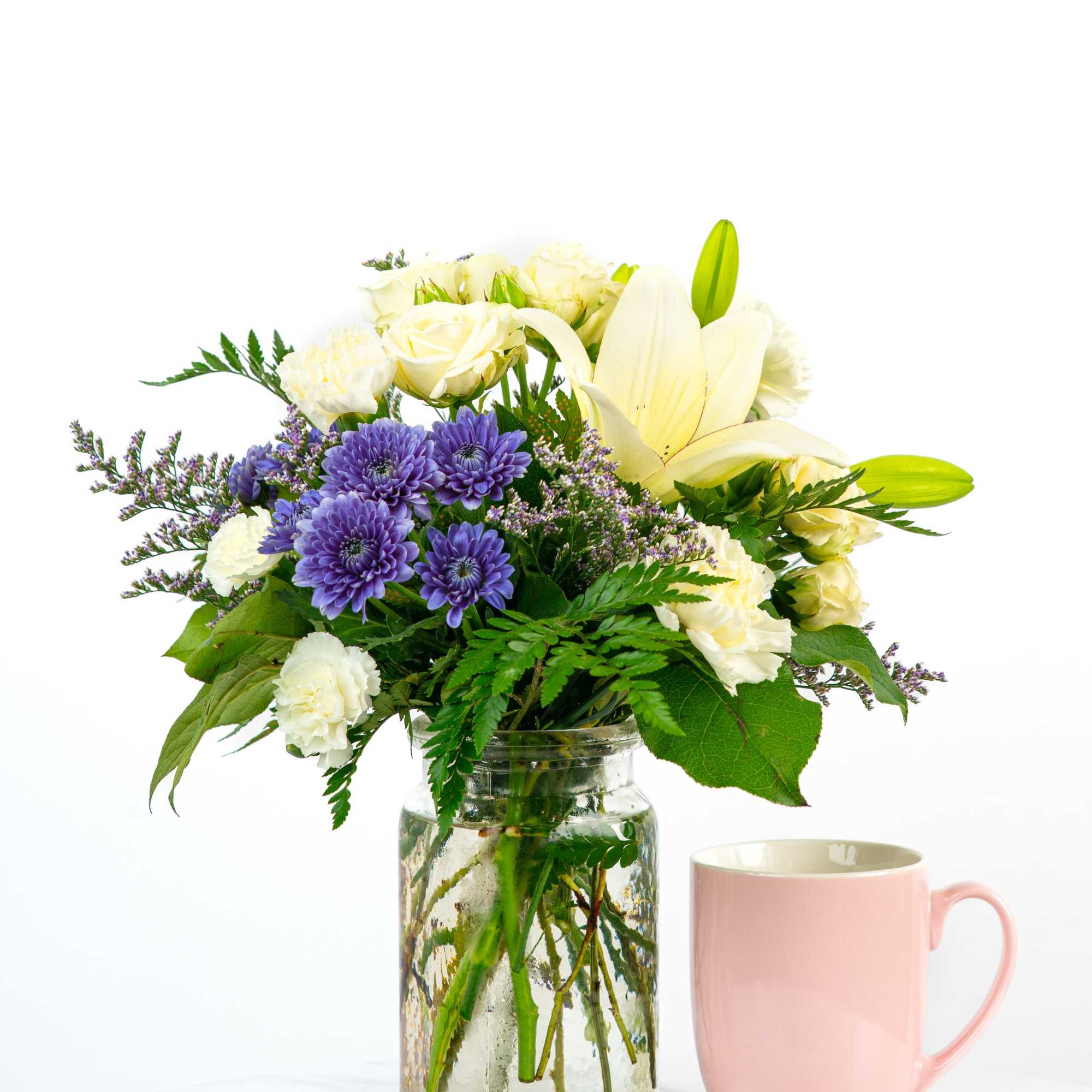 Bouquet of white lilies, roses, and purple flowers in a glass jar beside a pink mug