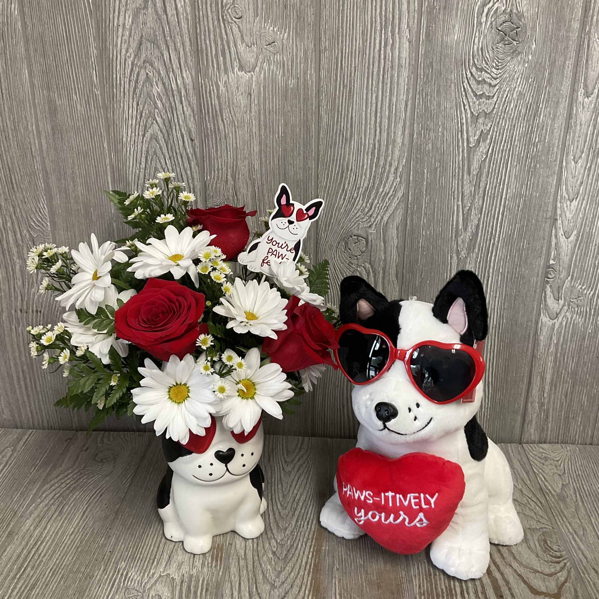 Red roses and white daisies in a dog-shaped vase beside a plush dog with heart sunglasses.