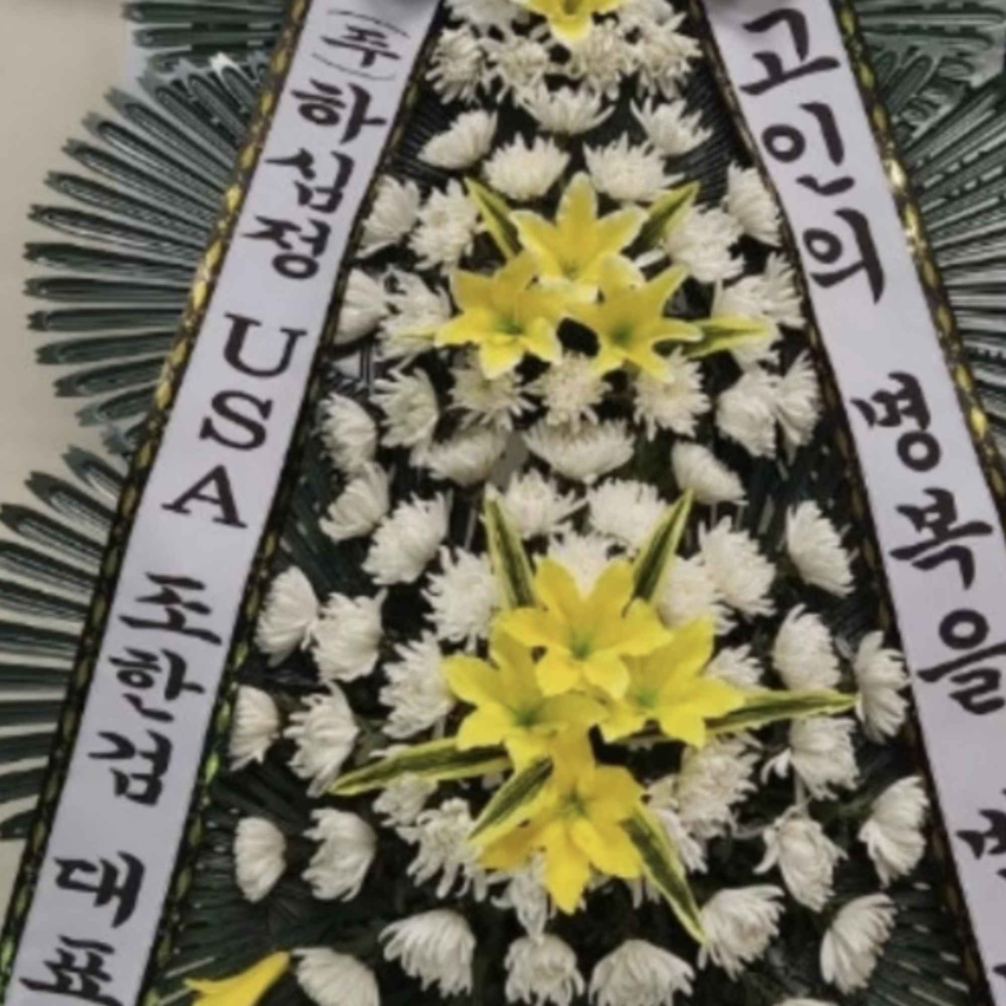 Large funeral wreath with yellow lilies and white flowers, framed by ribbons.
