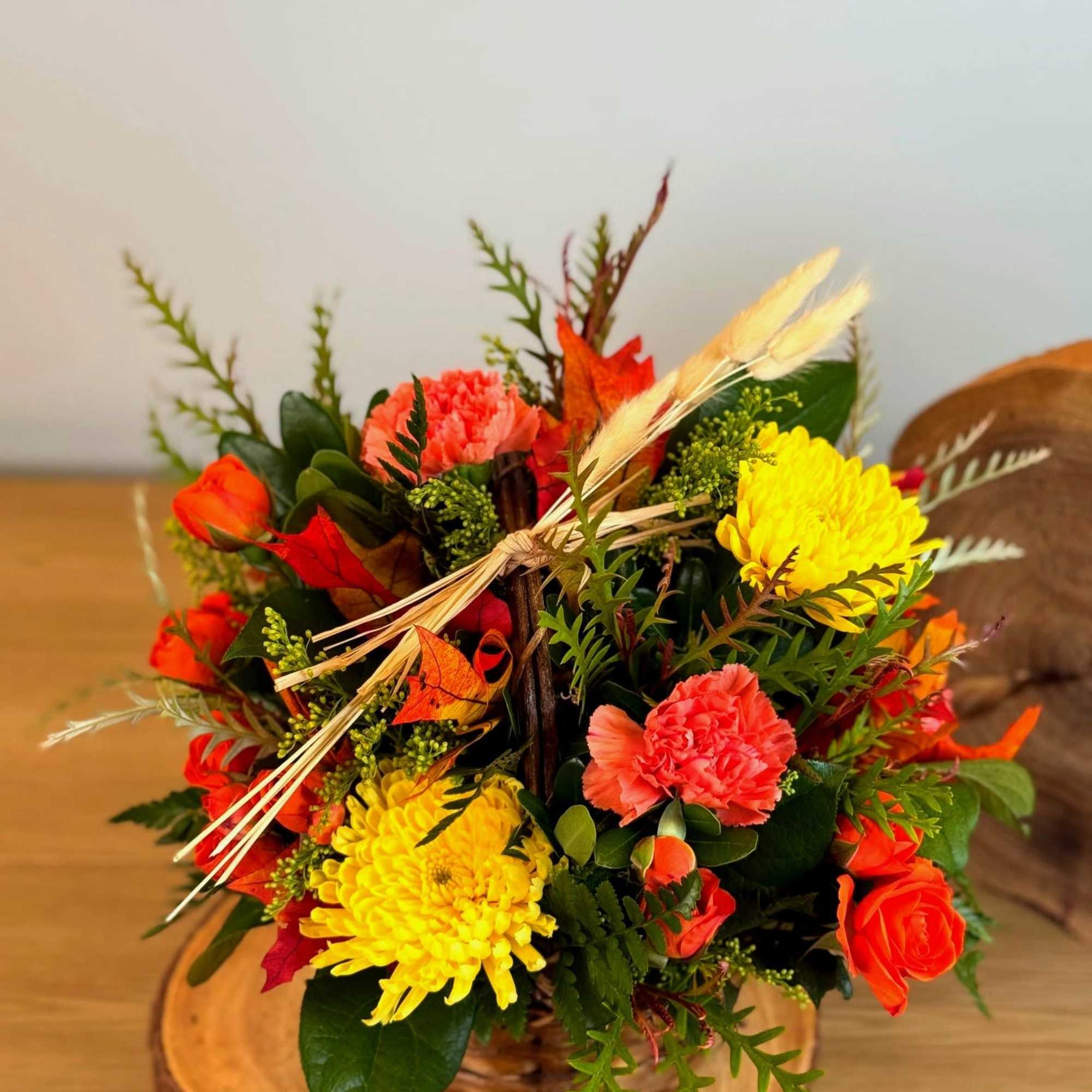 Autumn basket arrangement with yellow mums, orange roses and carnations accented with wheat on a wood slice base