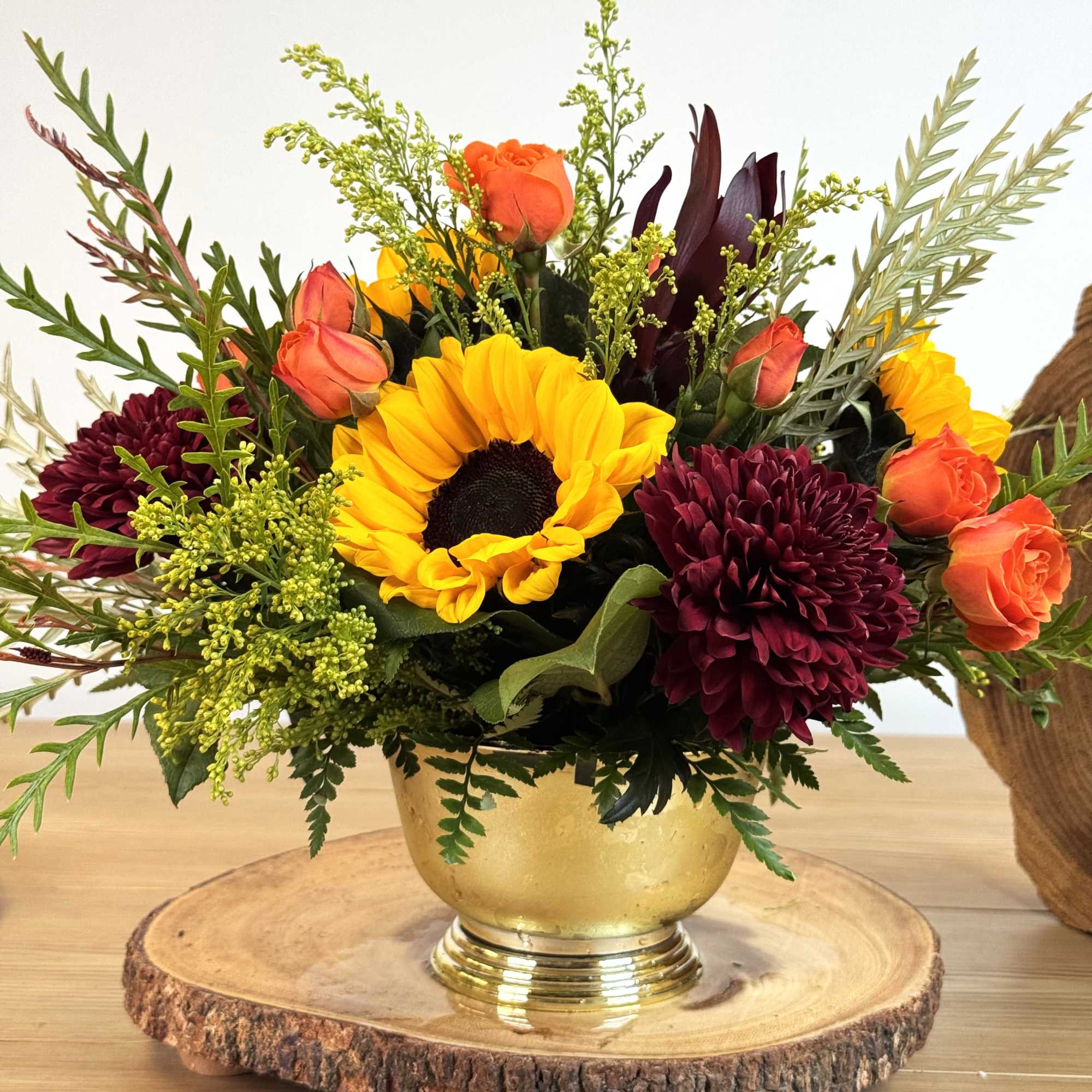 Low arrangement with a sunflower, orange roses, and burgundy mums in a gold bowl on a wood slice base