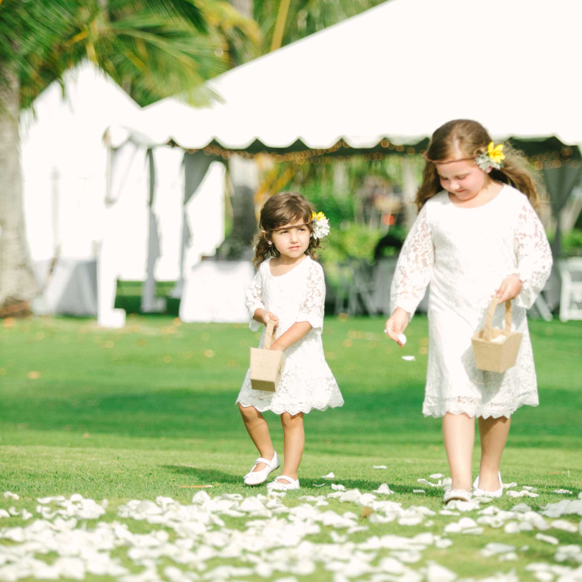 Two flower girls in white dresses scatter white petals on a lawn at an outdoor wedding