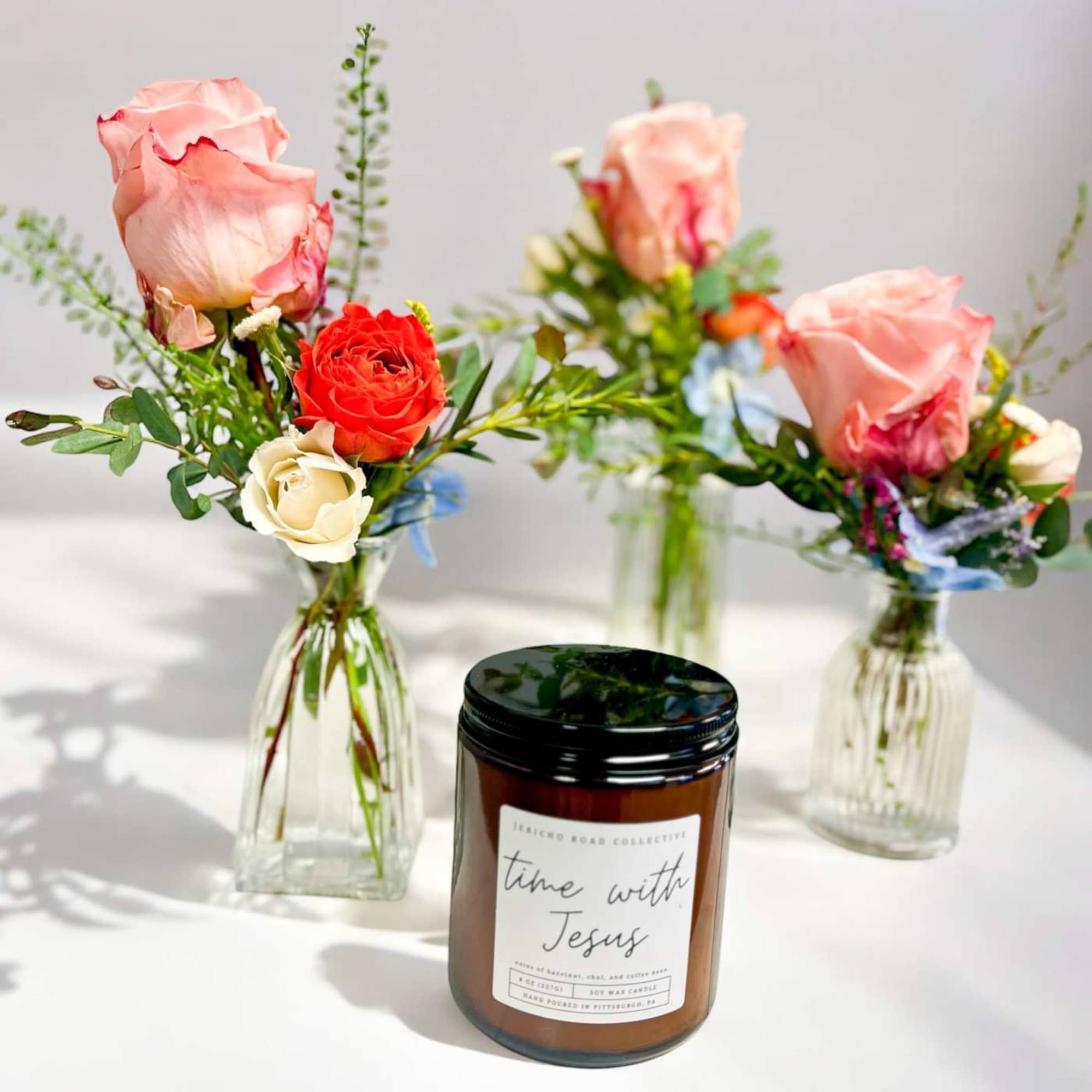 Three small bud vases with pink and red roses beside a brown jar candle on a white surface.