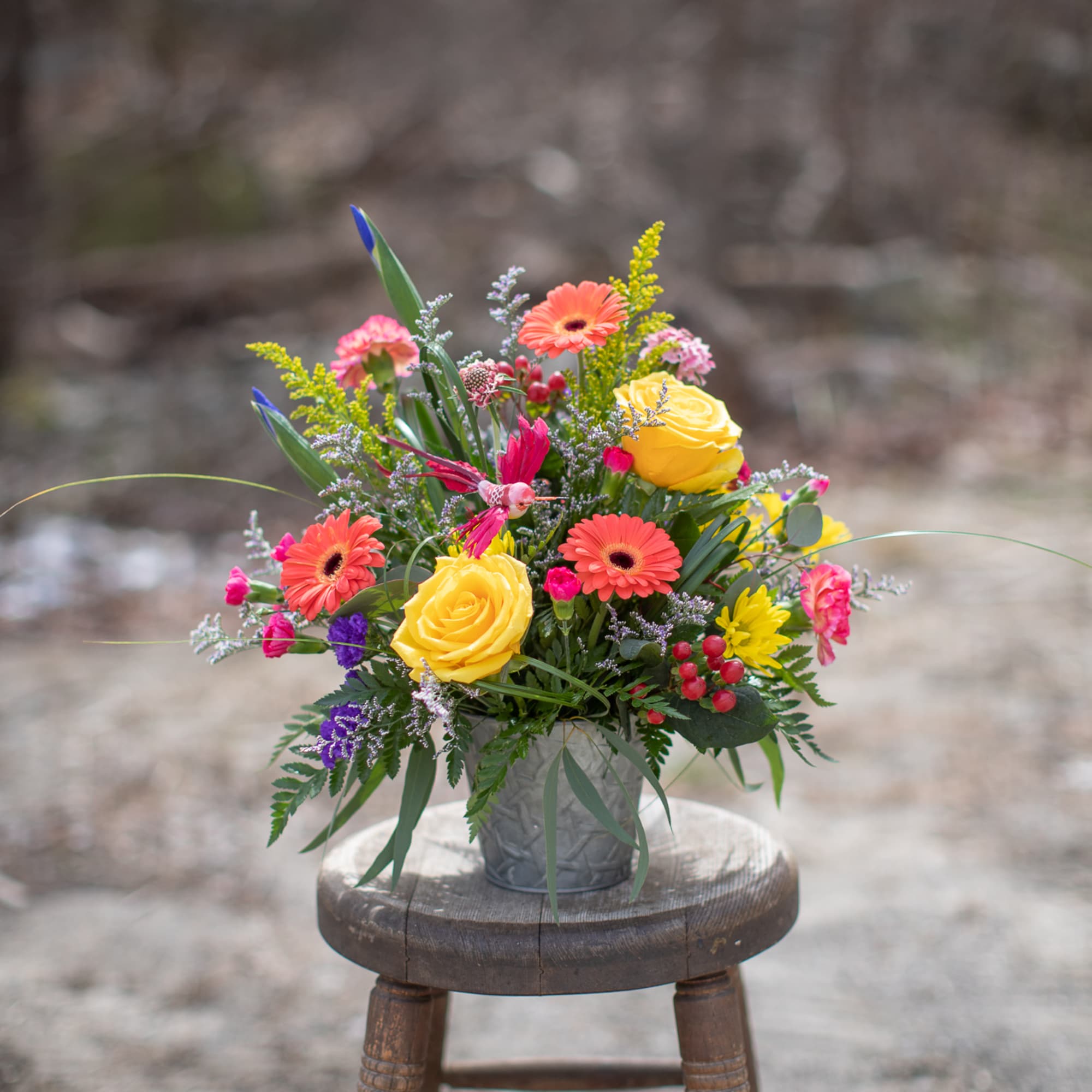 Colorful mixed flower arrangement in a small metal vase on a wooden stool