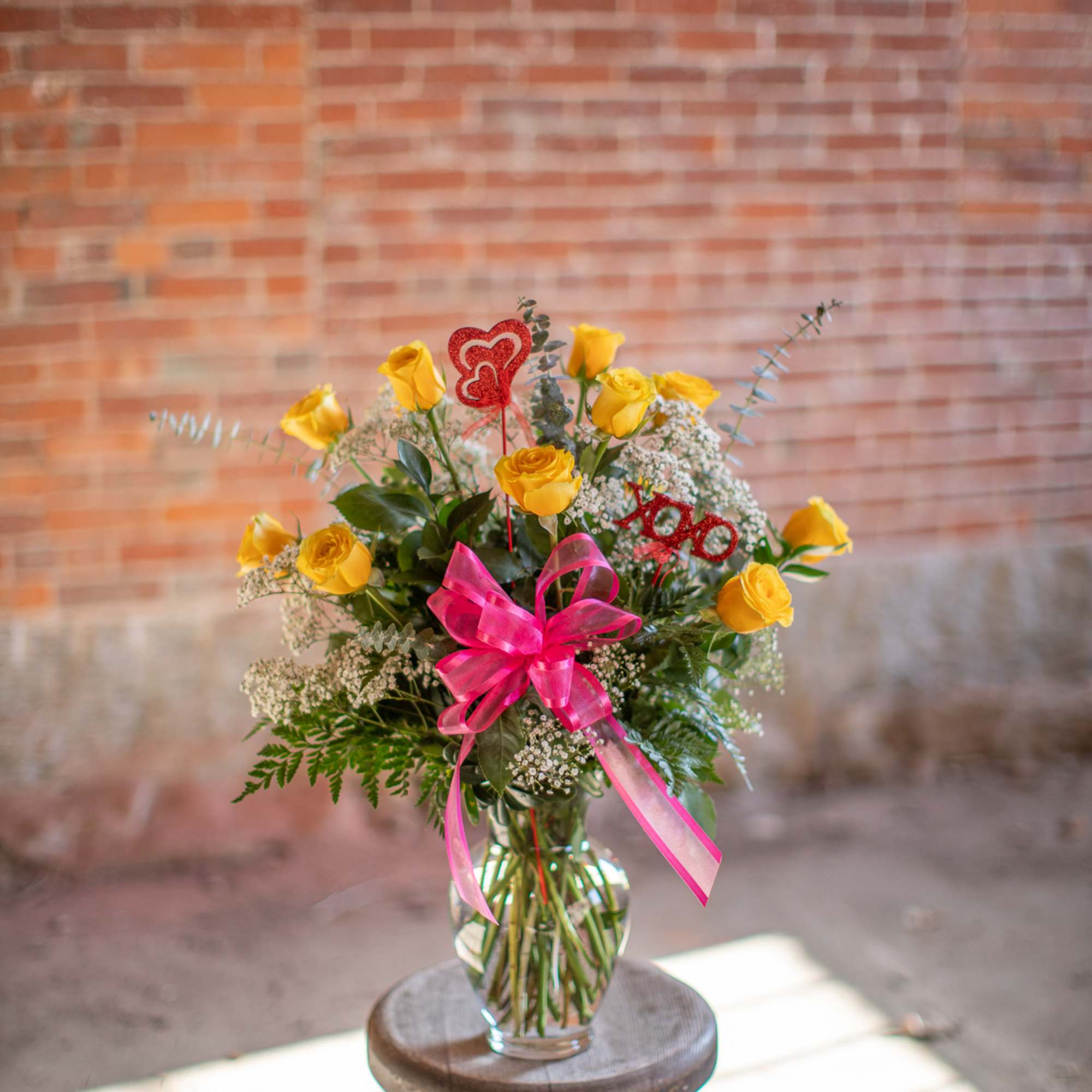 Yellow roses in a glass vase with pink ribbon and heart toppers