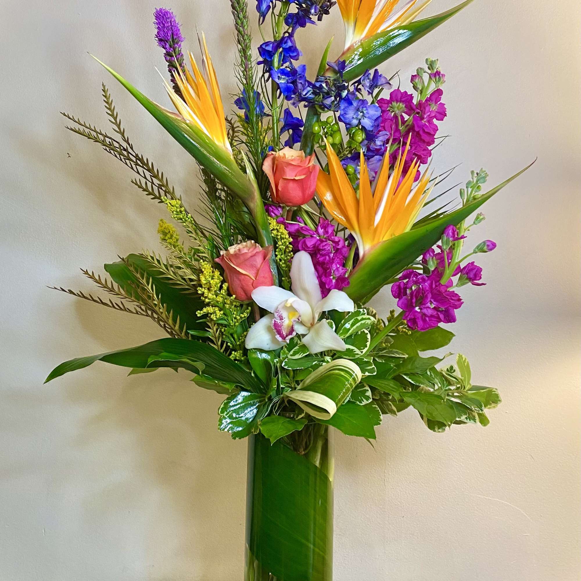 Tall tropical bouquet with orange bird-of-paradise, purple flowers, and pink roses in a glass vase