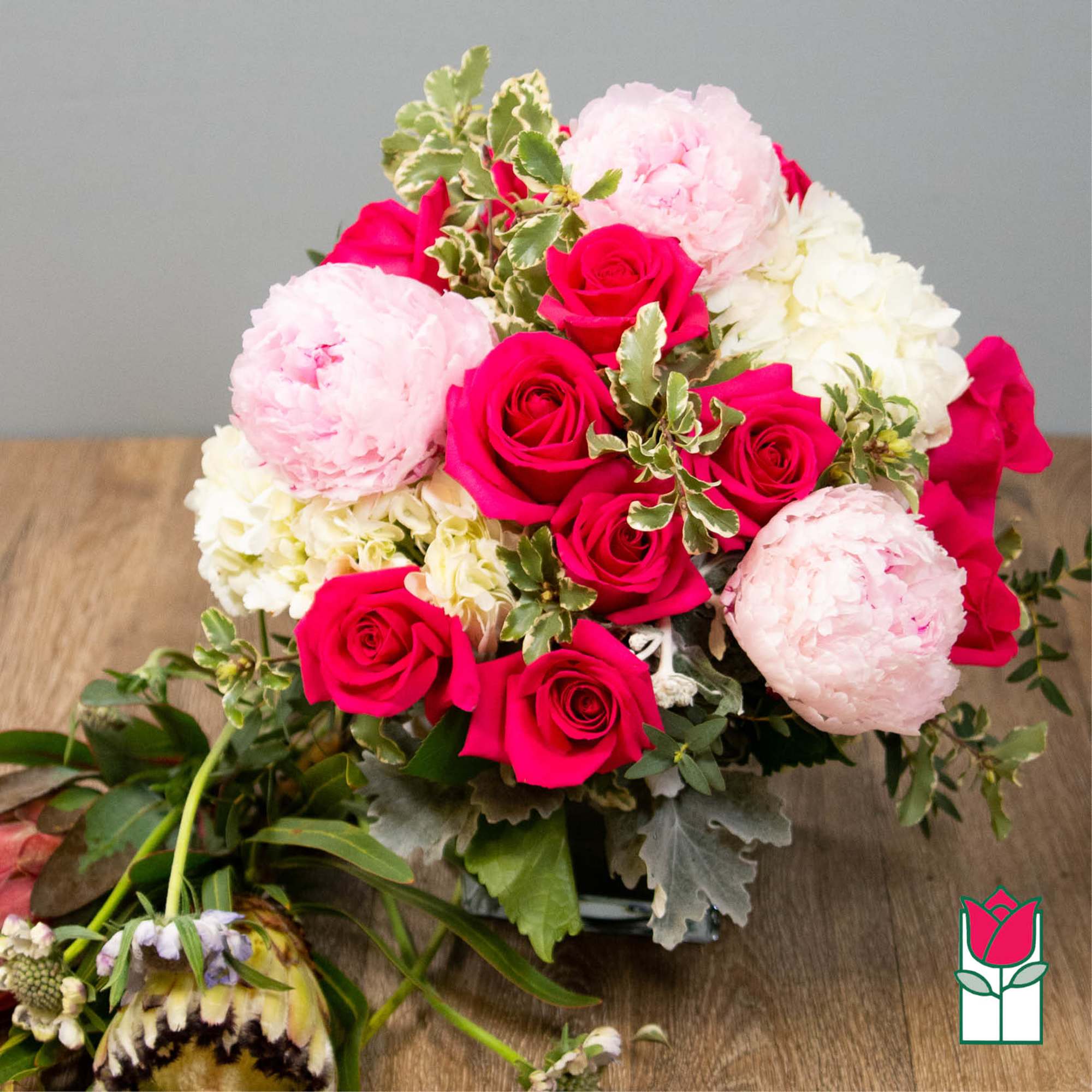 Compact arrangement of hot pink roses, pale pink peonies, and white blooms in a clear glass vase.