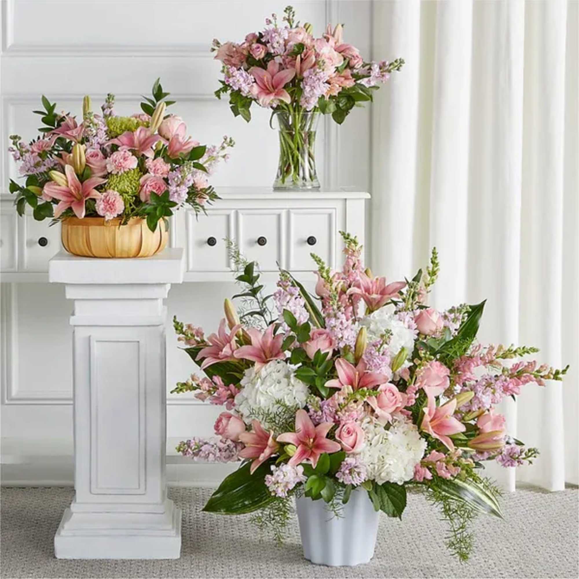 Set of pink and white floral arrangements in vases and a basket on white stands.
