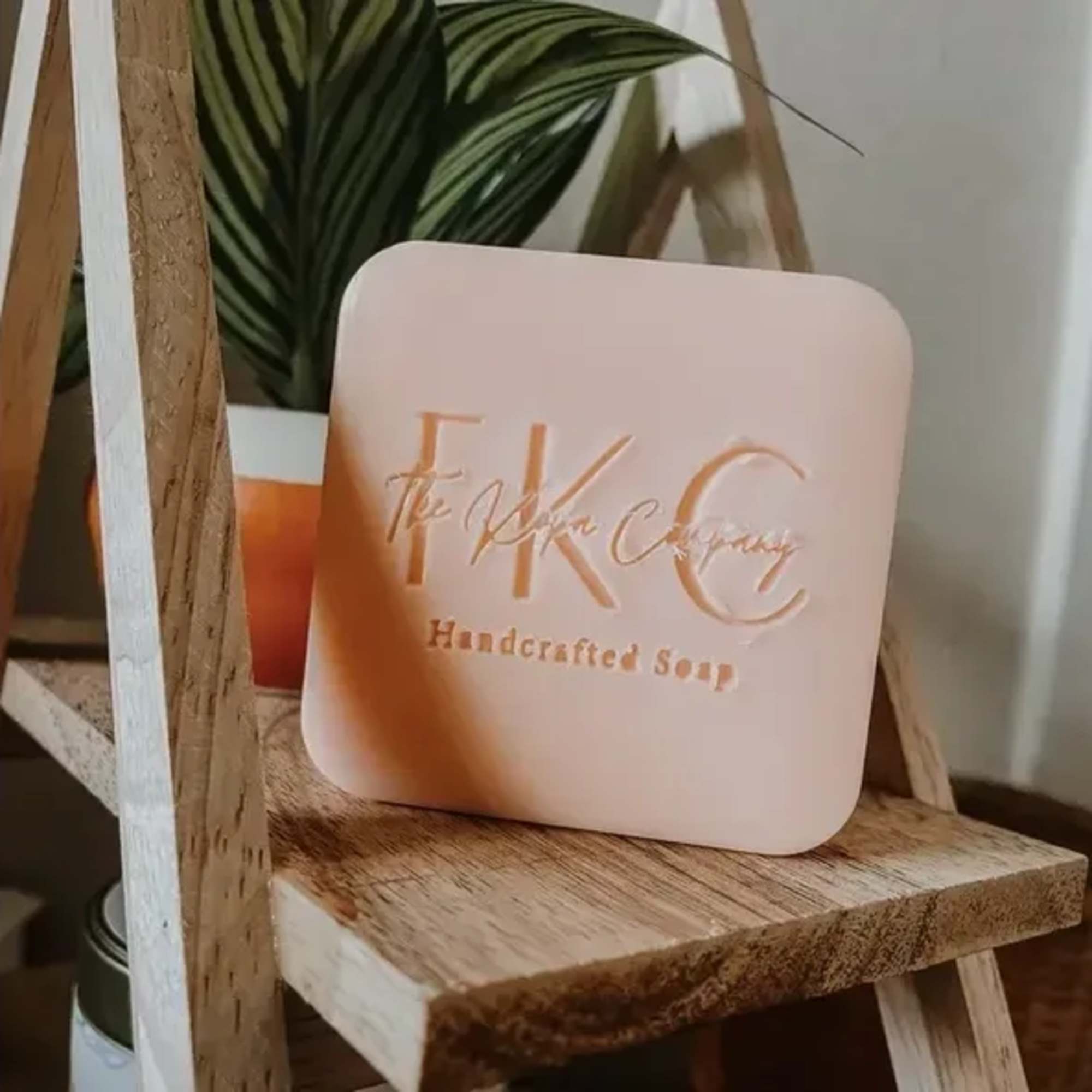 Square pink soap bar on a wooden shelf with a plant behind it