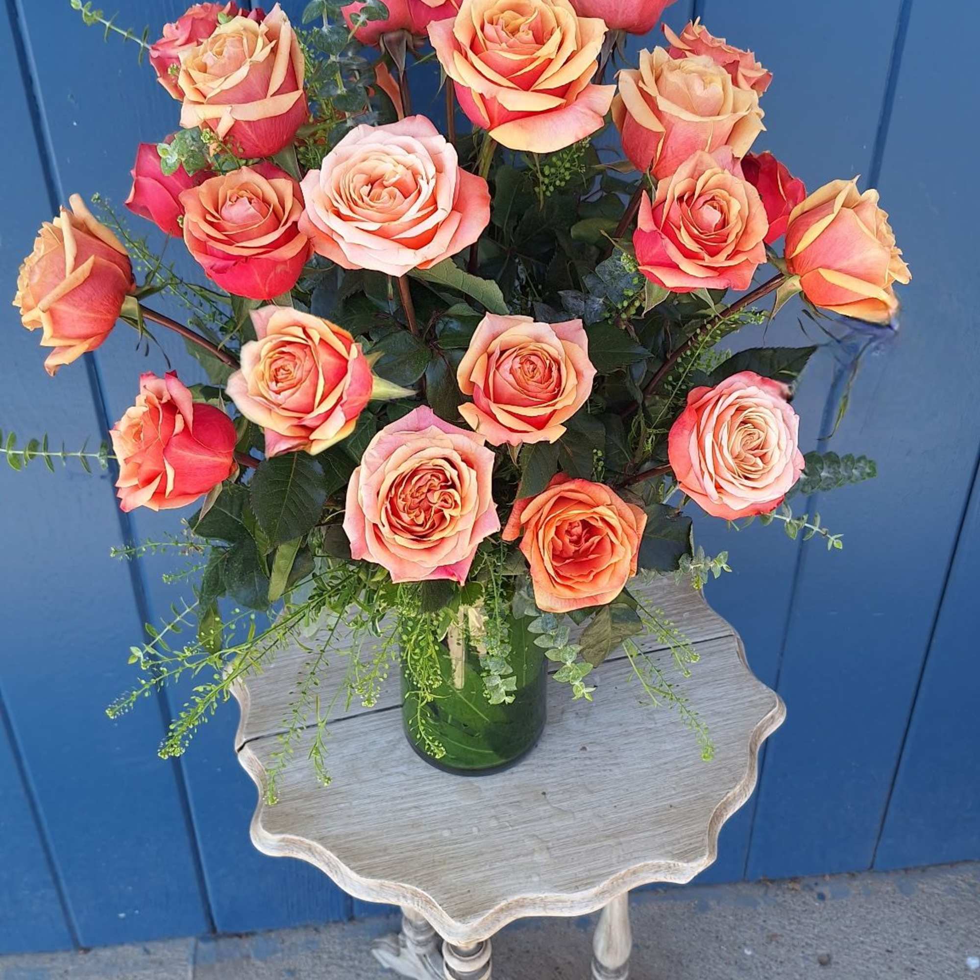 Tall arrangement of orange and peach roses in a green glass vase on a small wooden table.
