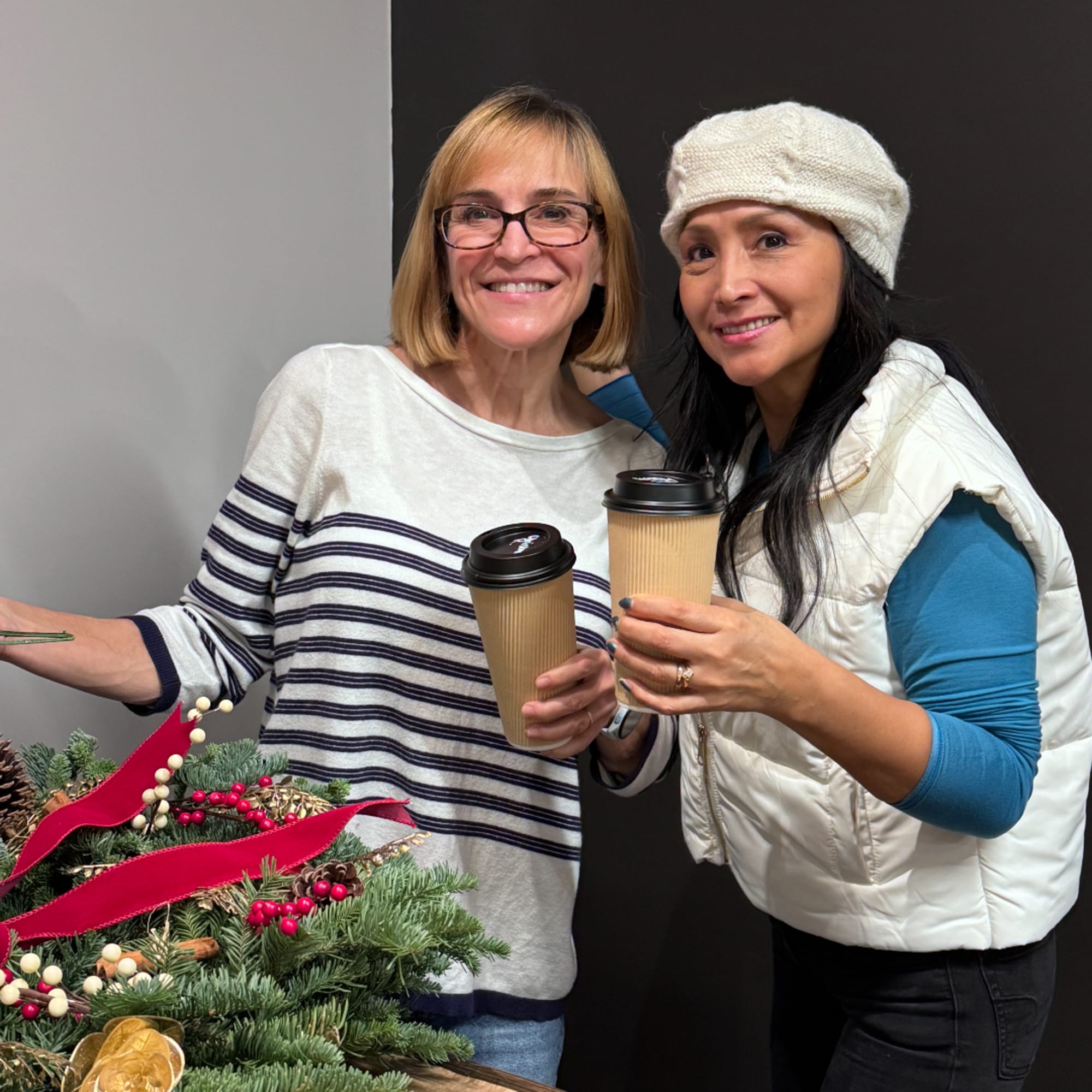 Two women holding coffee cups beside a decorated evergreen wreath