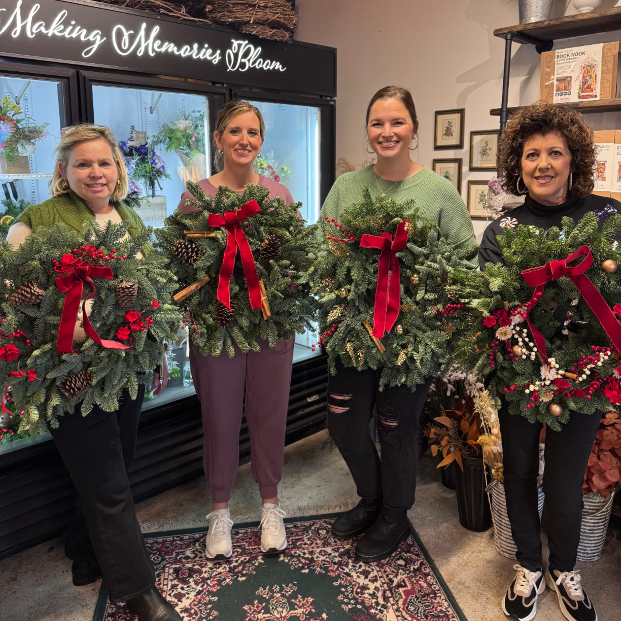 Four women hold large evergreen holiday wreaths with red bows.