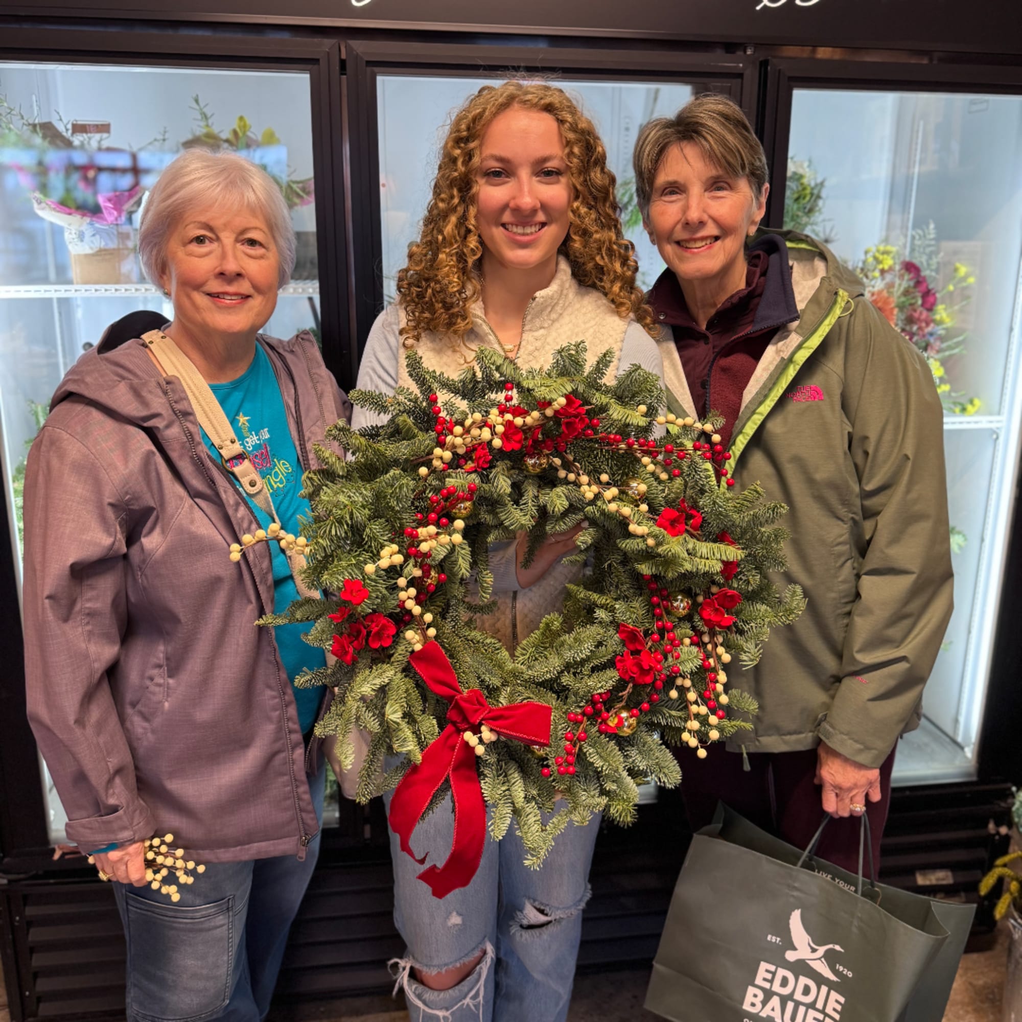 Three women hold a Christmas wreath with red berries and a red bow