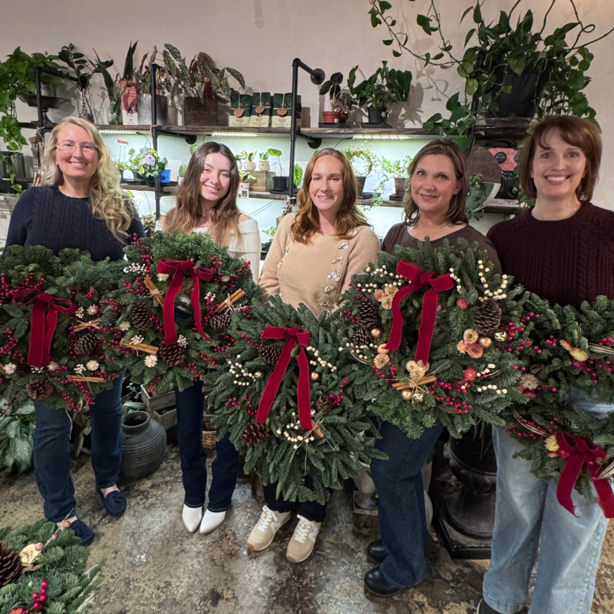 Five women hold large evergreen wreaths with red bows in a plant shop.